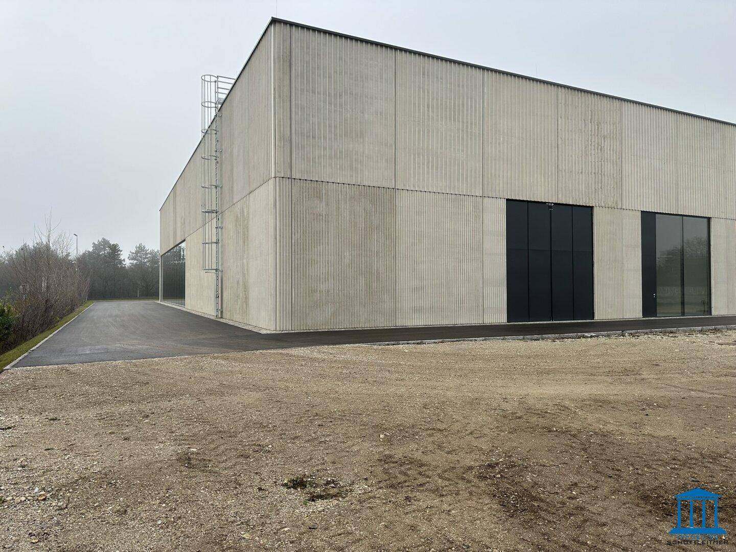 Ansicht eines modernen Industriegebäudes mit großen Toren und einer unbefestigten Fläche davor.