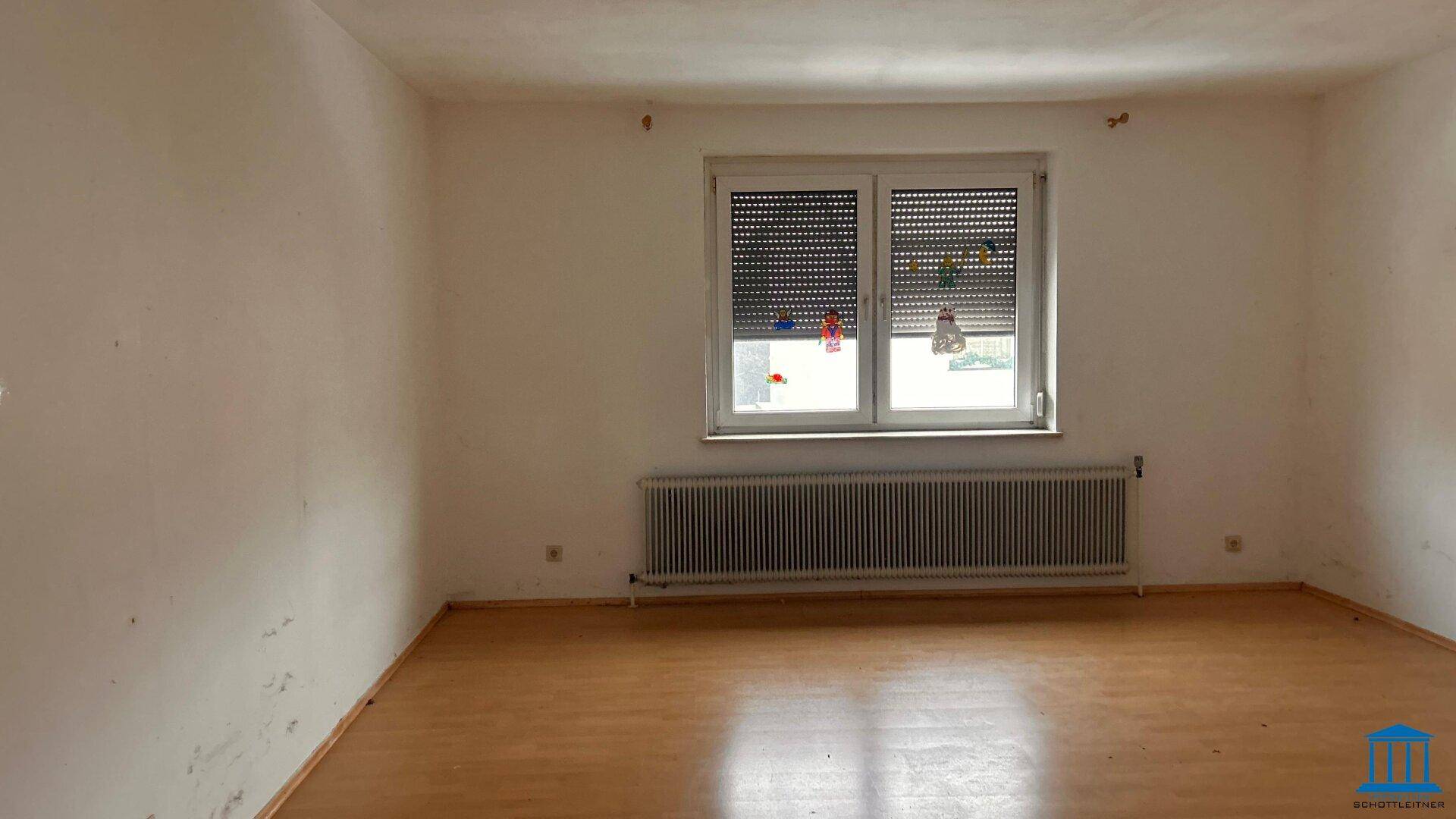 Leerer Raum mit hellem Holzboden, großem Fenster und Heizkörper unter der Fensterbank.