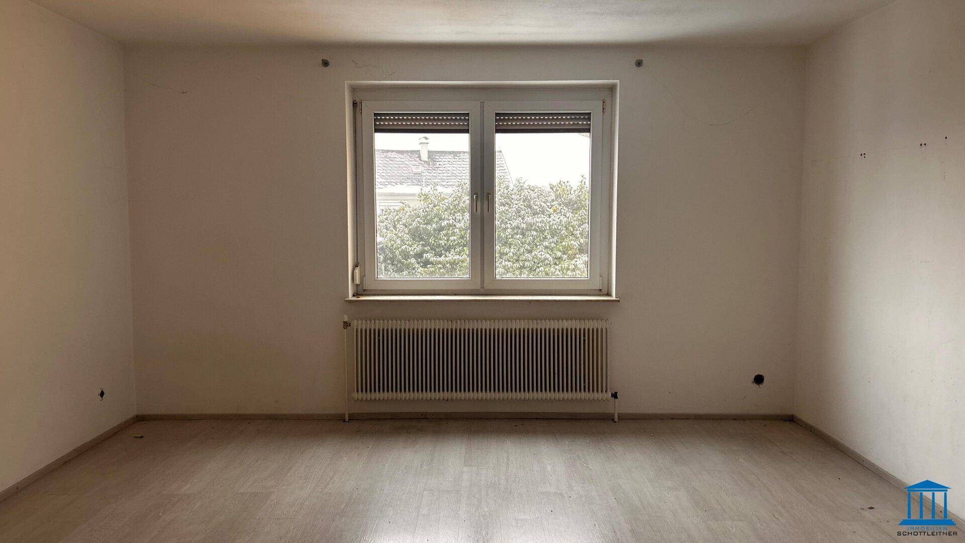 Heller, leerer Raum mit Holzboden und Fenster, das einen Blick ins Grüne bietet.