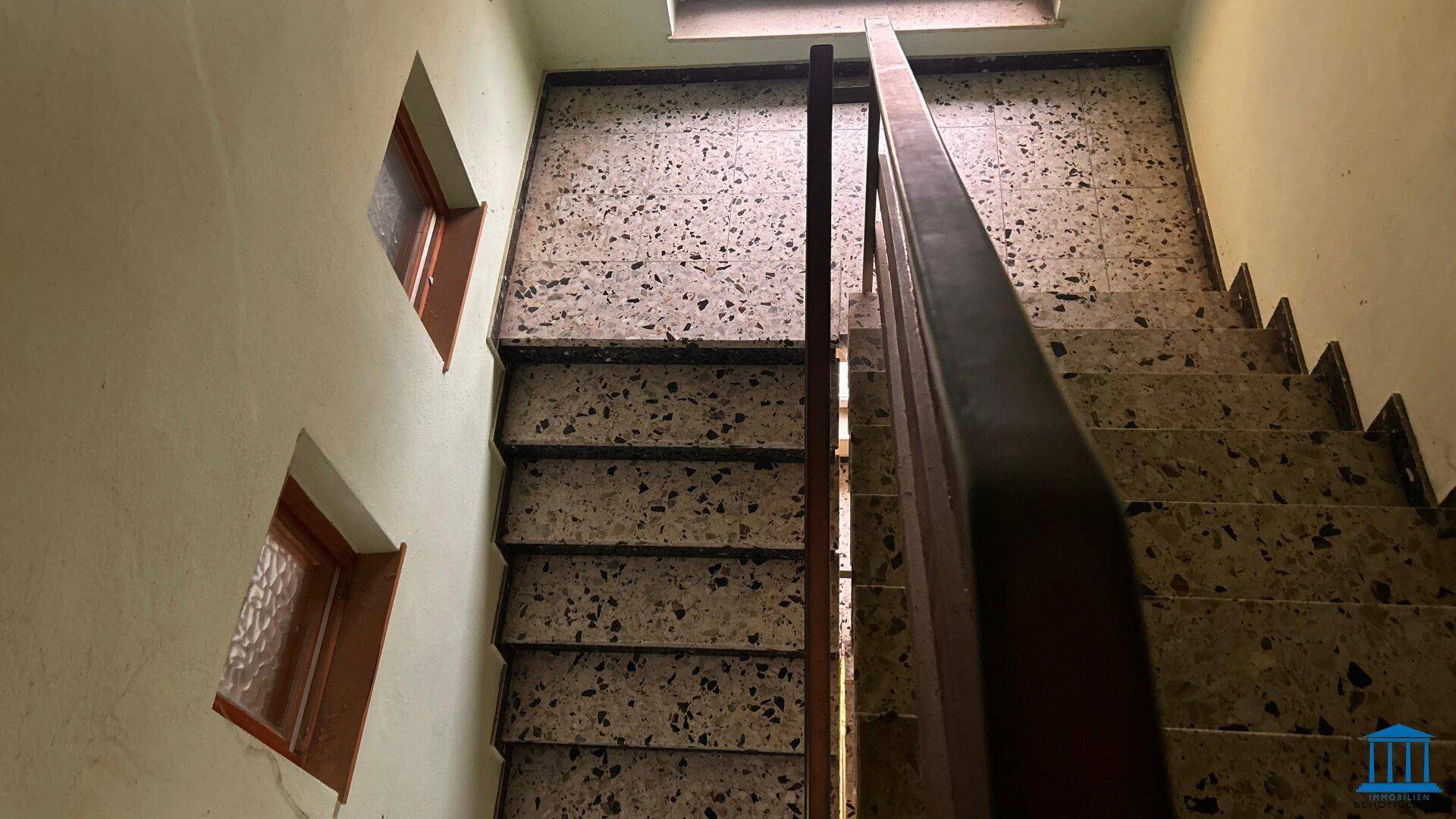 Treppenhaus mit Terrazzoboden und Holzgeländer, das zu den oberen Etagen führt.