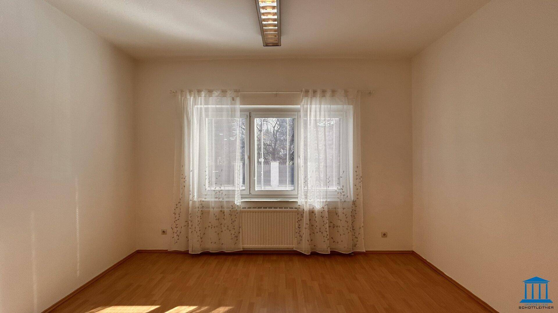 Heller Büroraum mit Fenster, Vorhängen und Holzfußboden, gut beleuchtet durch Tageslicht.