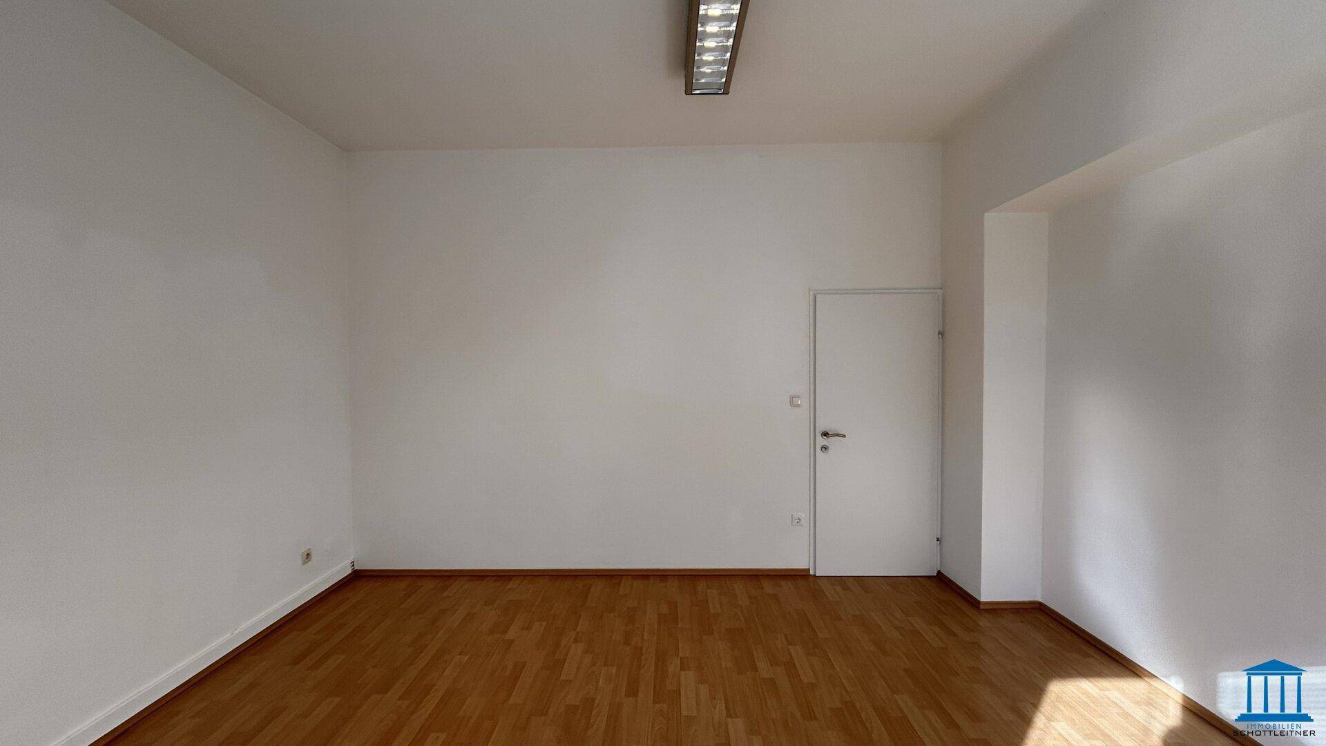 Leerer Büroraum mit weiß gestrichenen Wänden, Holzboden und einer einfachen Tür.