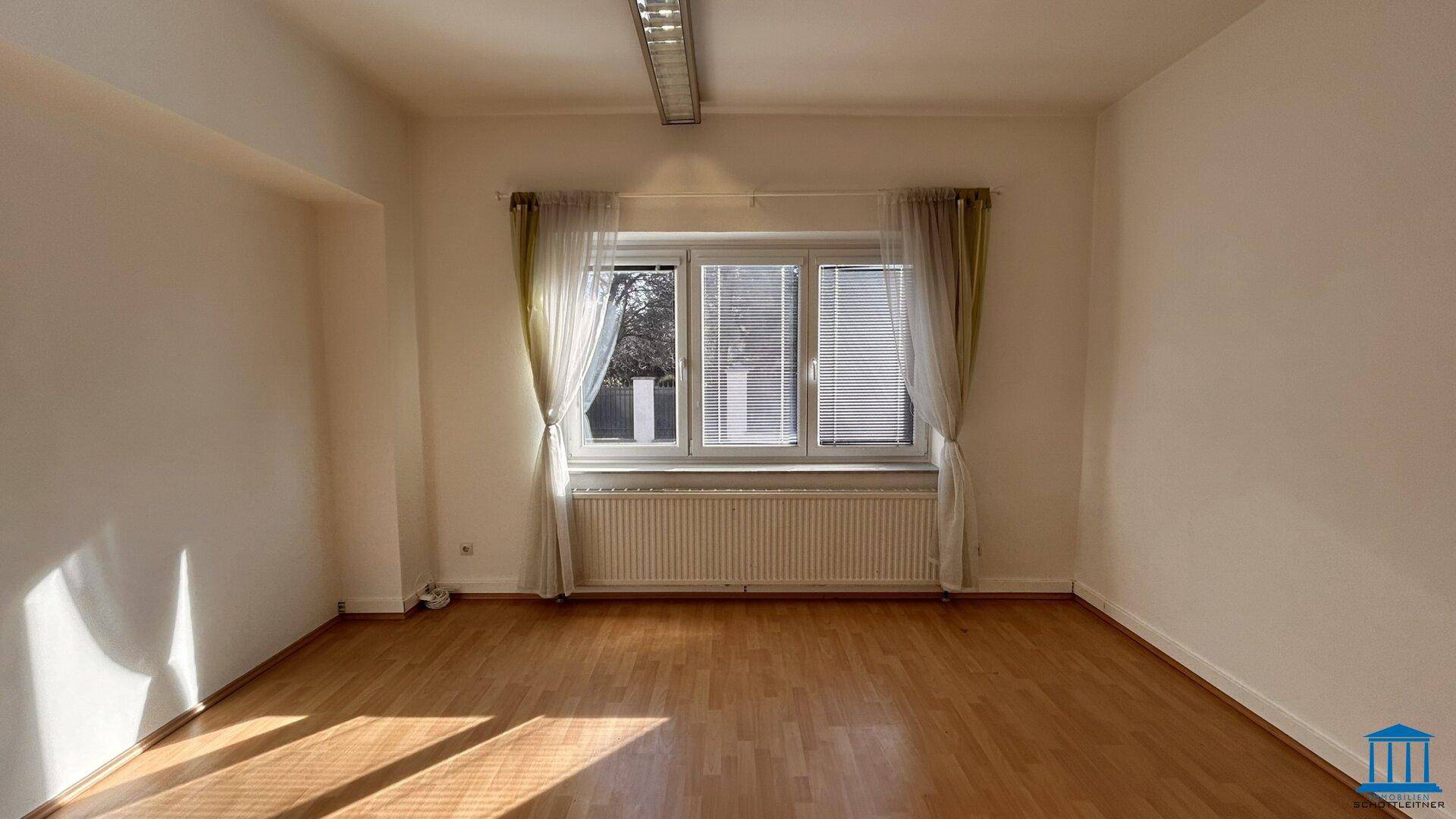 Heller Büroraum mit Fenster, Vorhängen und Holzfußboden, gut beleuchtet durch Tageslicht.