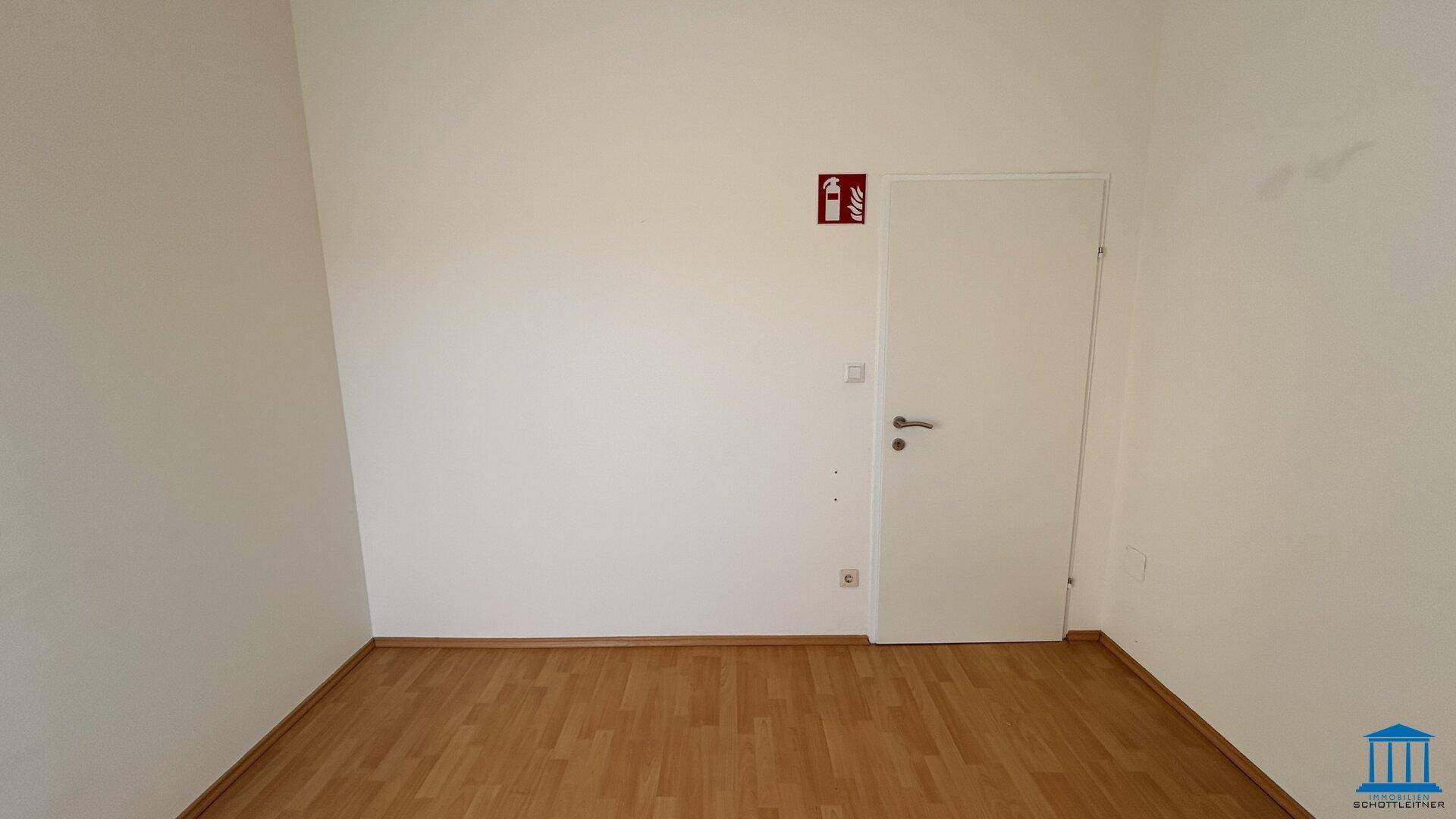 Leerer Büroraum mit weiß gestrichenen Wänden, Holzboden und einer Tür mit Feuerlöscher-Symbol.