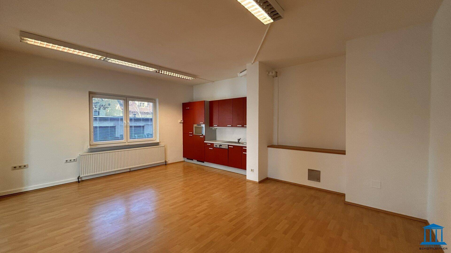Heller Büroraum mit roter Küchenzeile, Holzfußboden und großem Fenster.