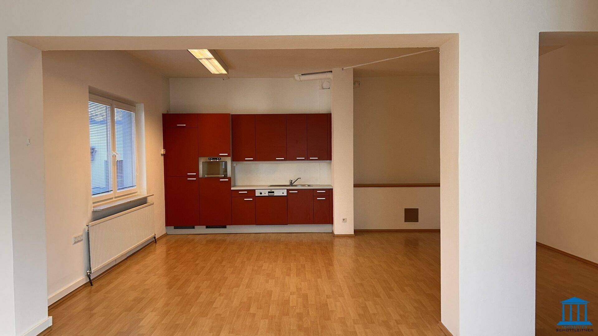 Offener Bürobereich mit integrierter roter Küchenzeile und Blick auf einen weiteren Raum.
