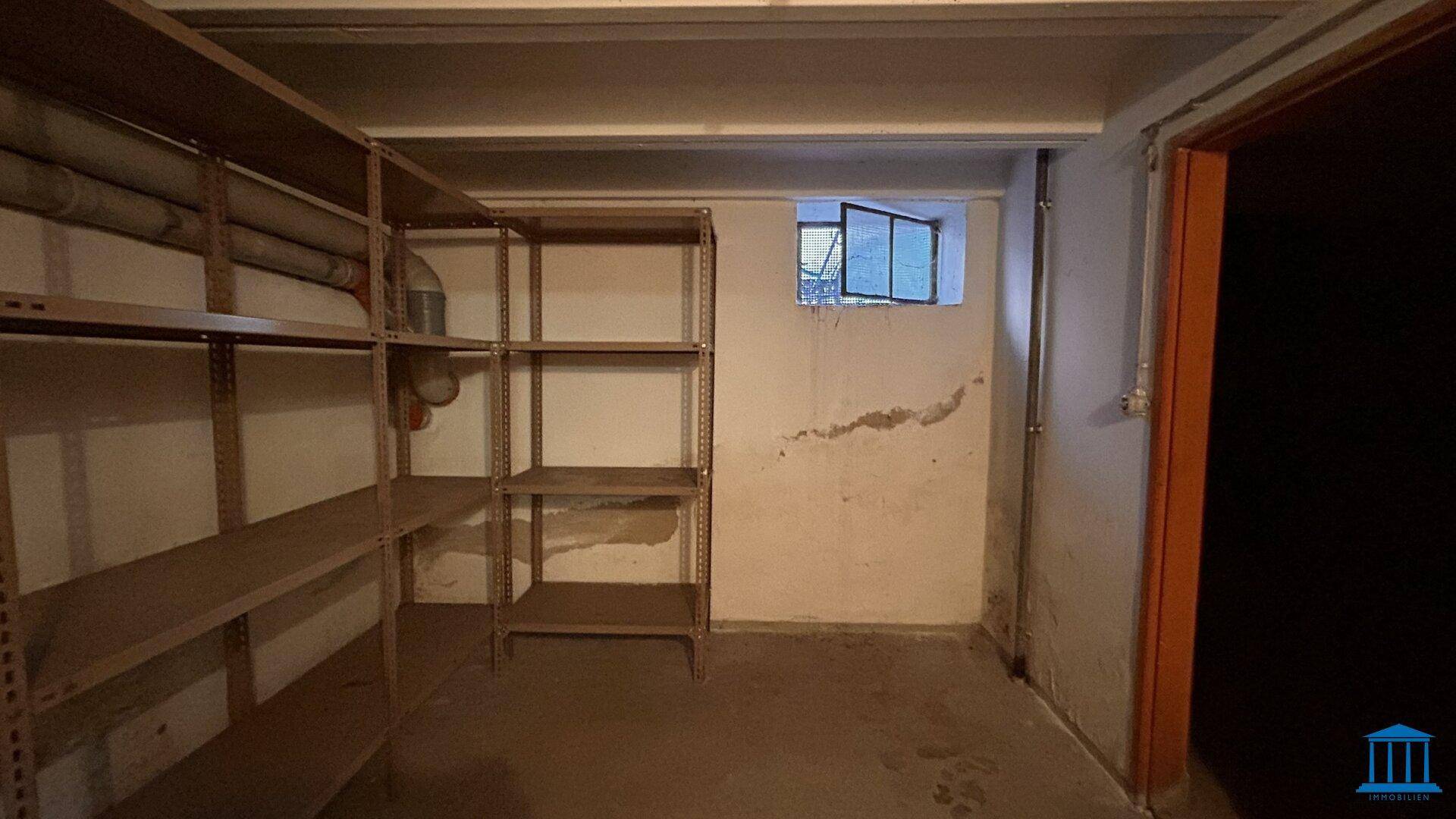 Kellerraum mit Regalen, kleinem Fenster und sichtbaren Rohren an der Decke.
