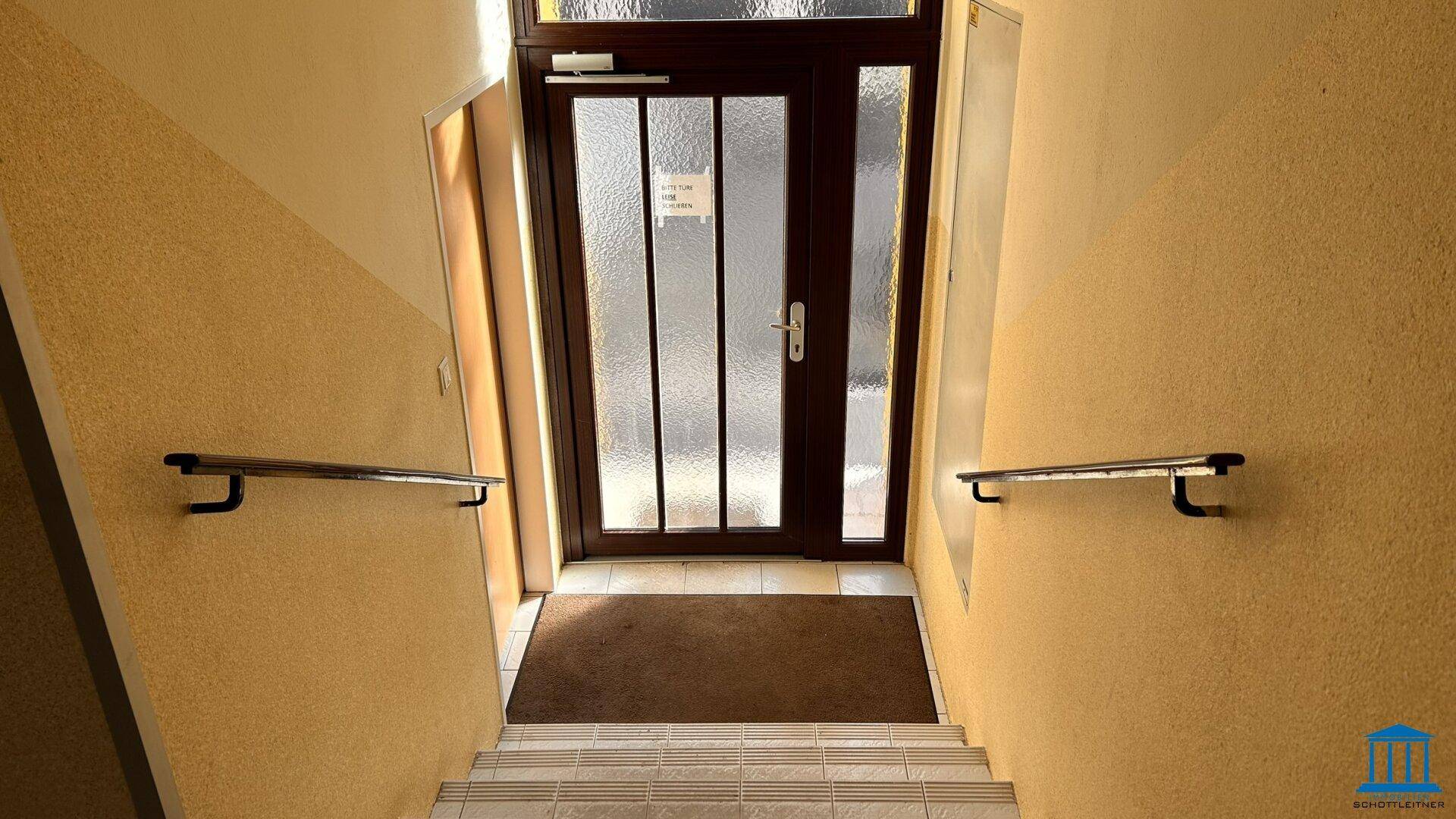 Eingangsbereich mit Treppe, Handläufen und einer Glastür zum Gebäude.