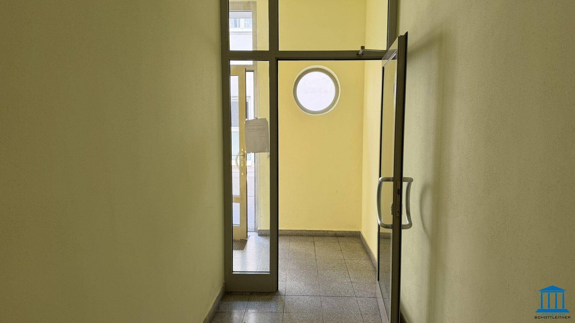 Heller Eingangsbereich mit Glastür und runden Fenstern, bietet Zugang zum Gebäude.