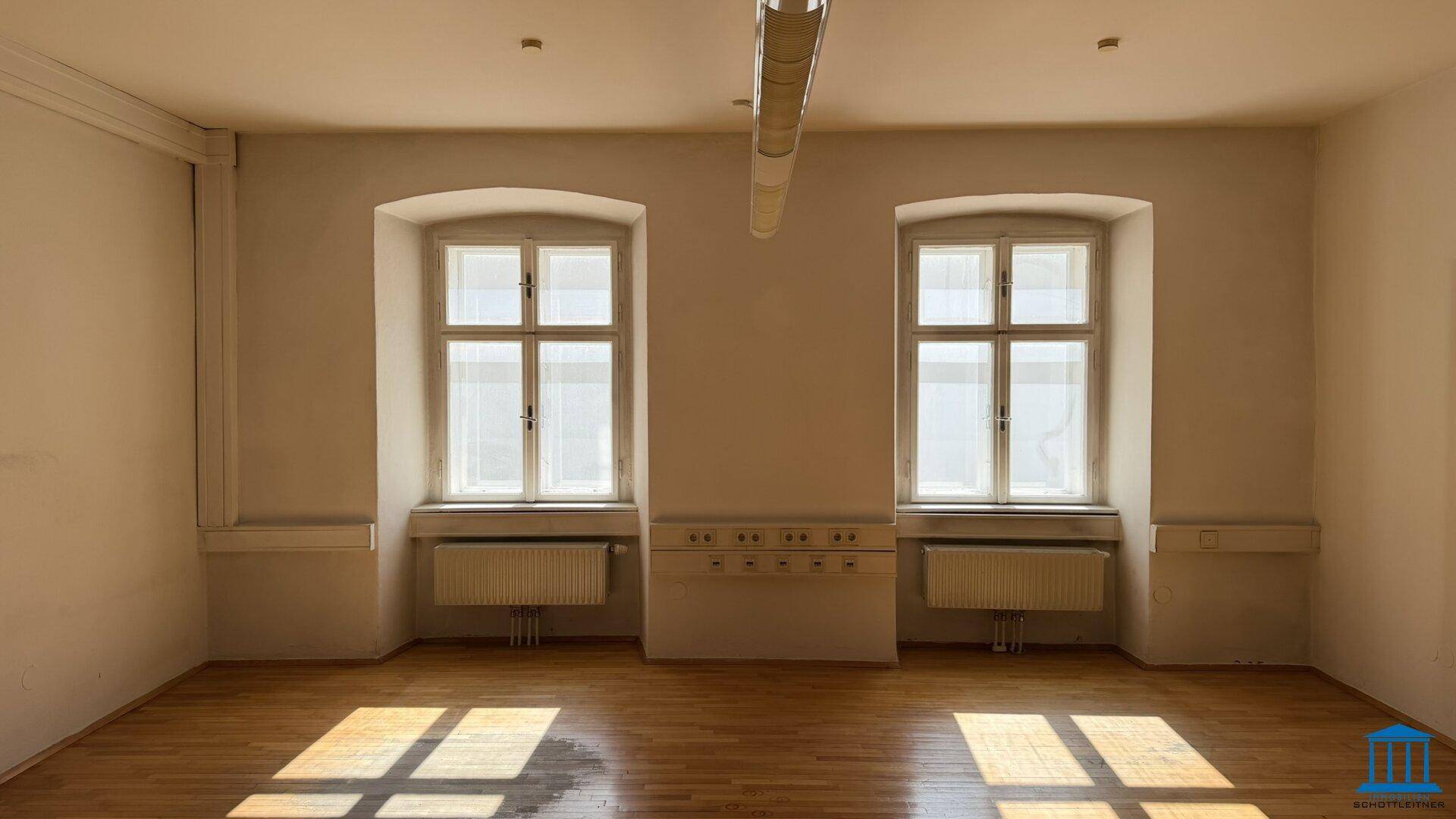 Geräumiger Büroraum mit zwei großen Fenstern und Holzboden, bietet viel Tageslicht.