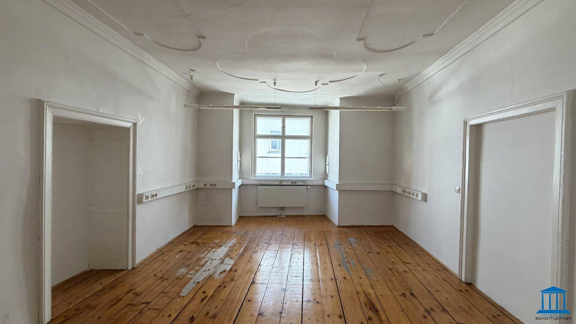 Heller Büroraum mit Holzdielenboden, Stuckdecke und einem Fenster, das Tageslicht hereinlässt.