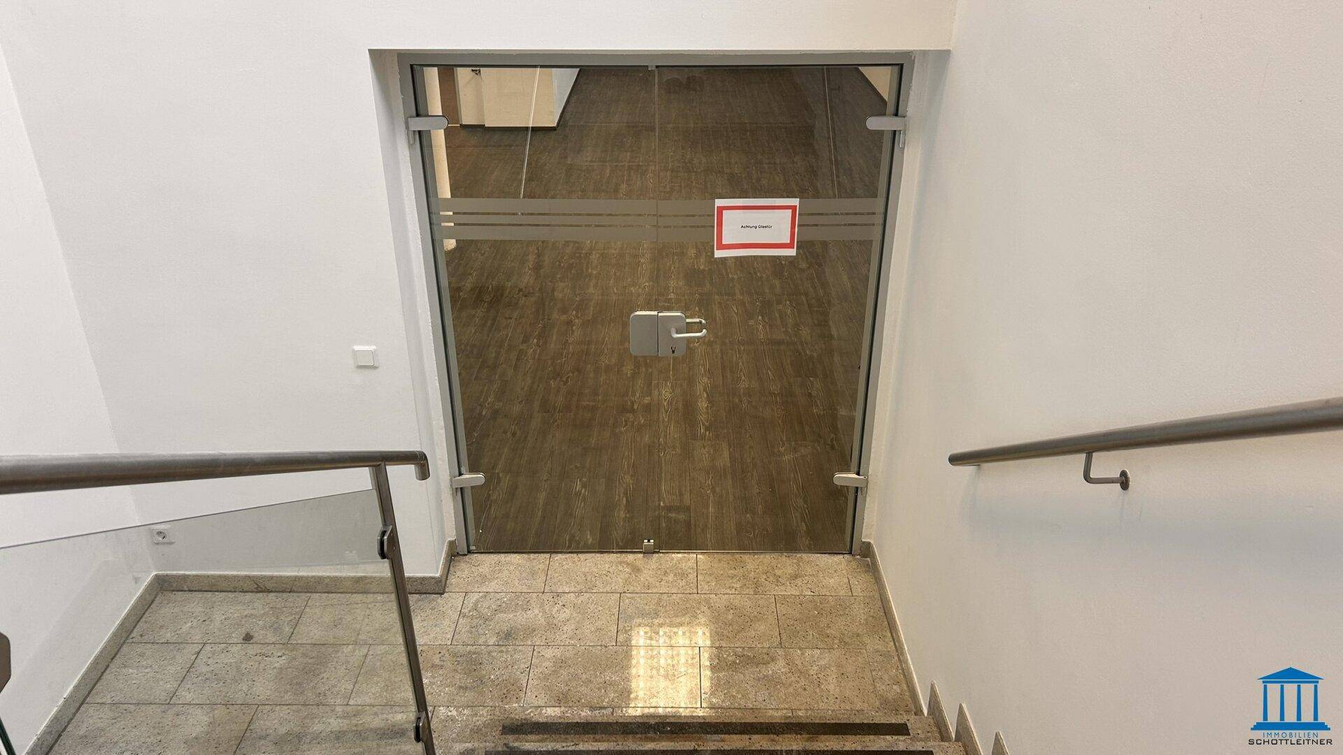 Treppenhaus mit Granitstufen und Glasgeländer, das zu einer Glastür führt.