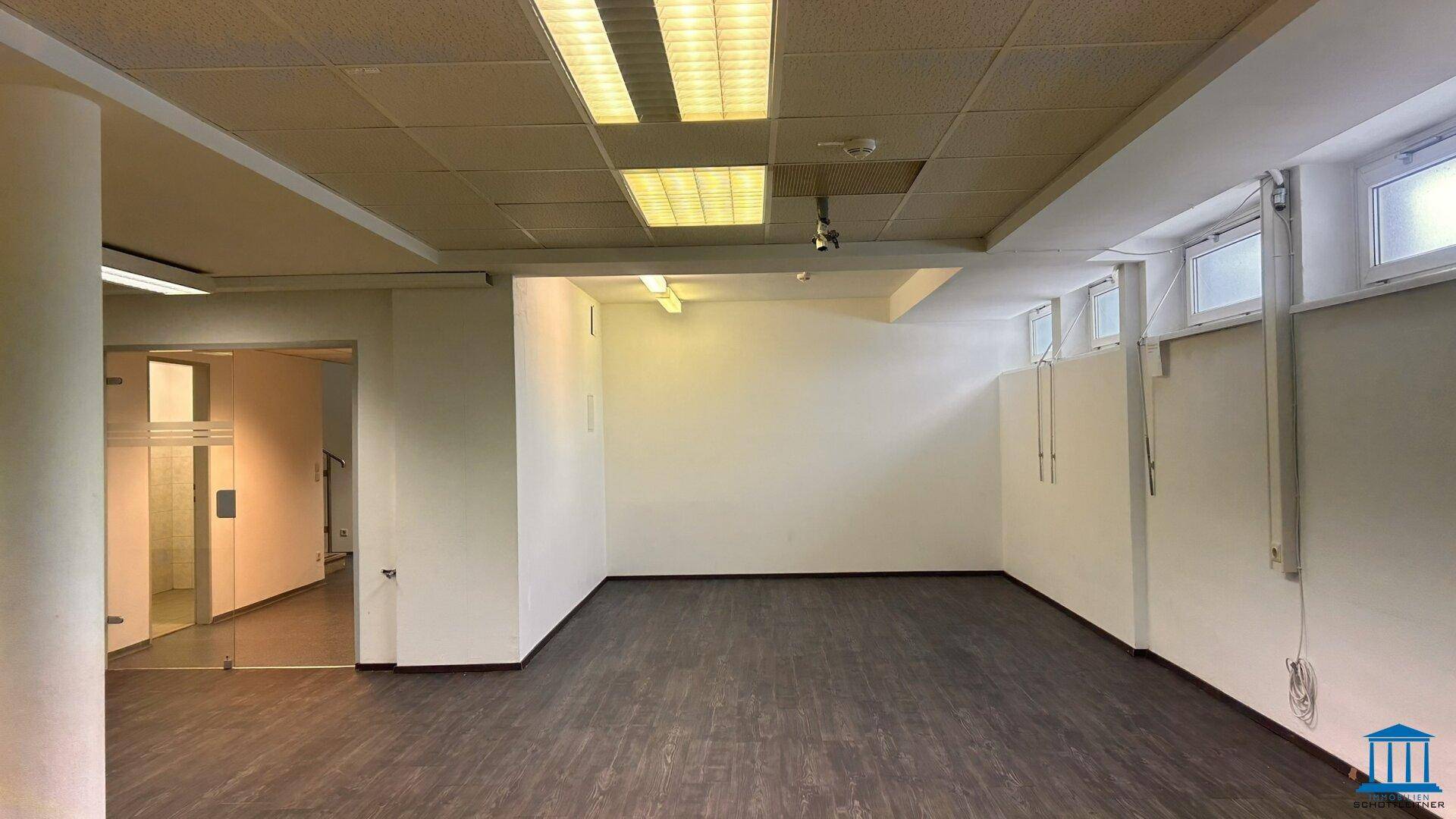 Geräumiger Büroraum mit dunklem Boden, Deckenbeleuchtung und Zugang durch eine Glastür.