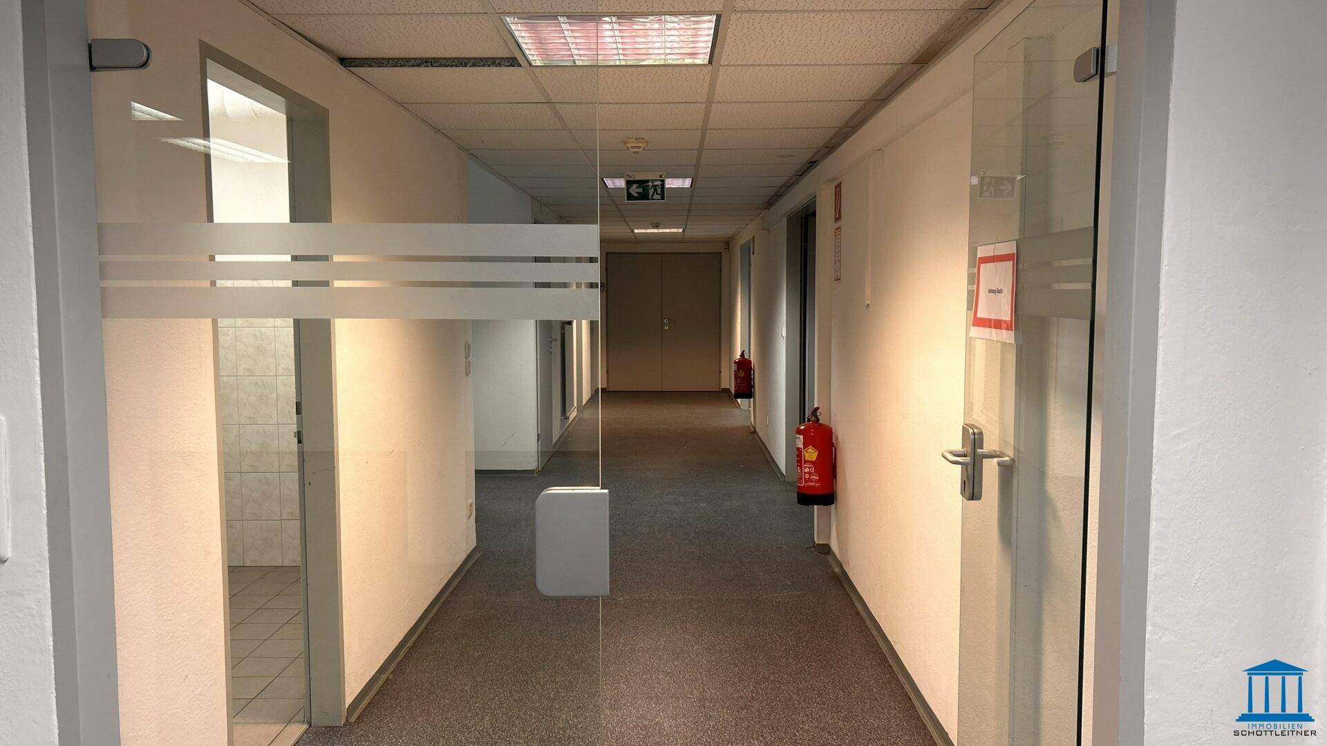 Langer Flur mit Teppichboden, Glastüren zu Büros und Brandschutzvorrichtungen.