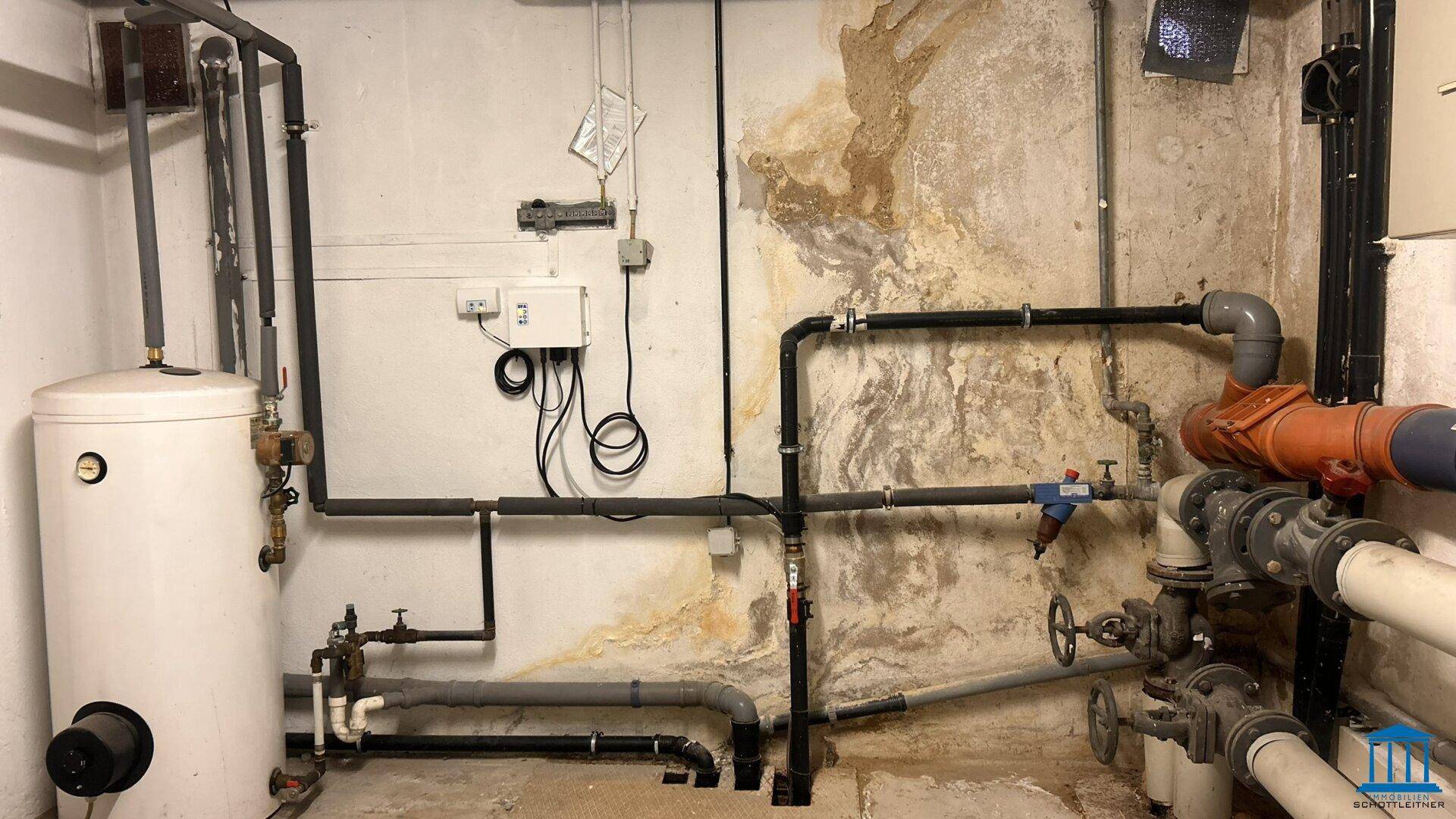 Technikraum mit Warmwasserspeicher, zahlreichen Rohren und Installationen an der Wand.