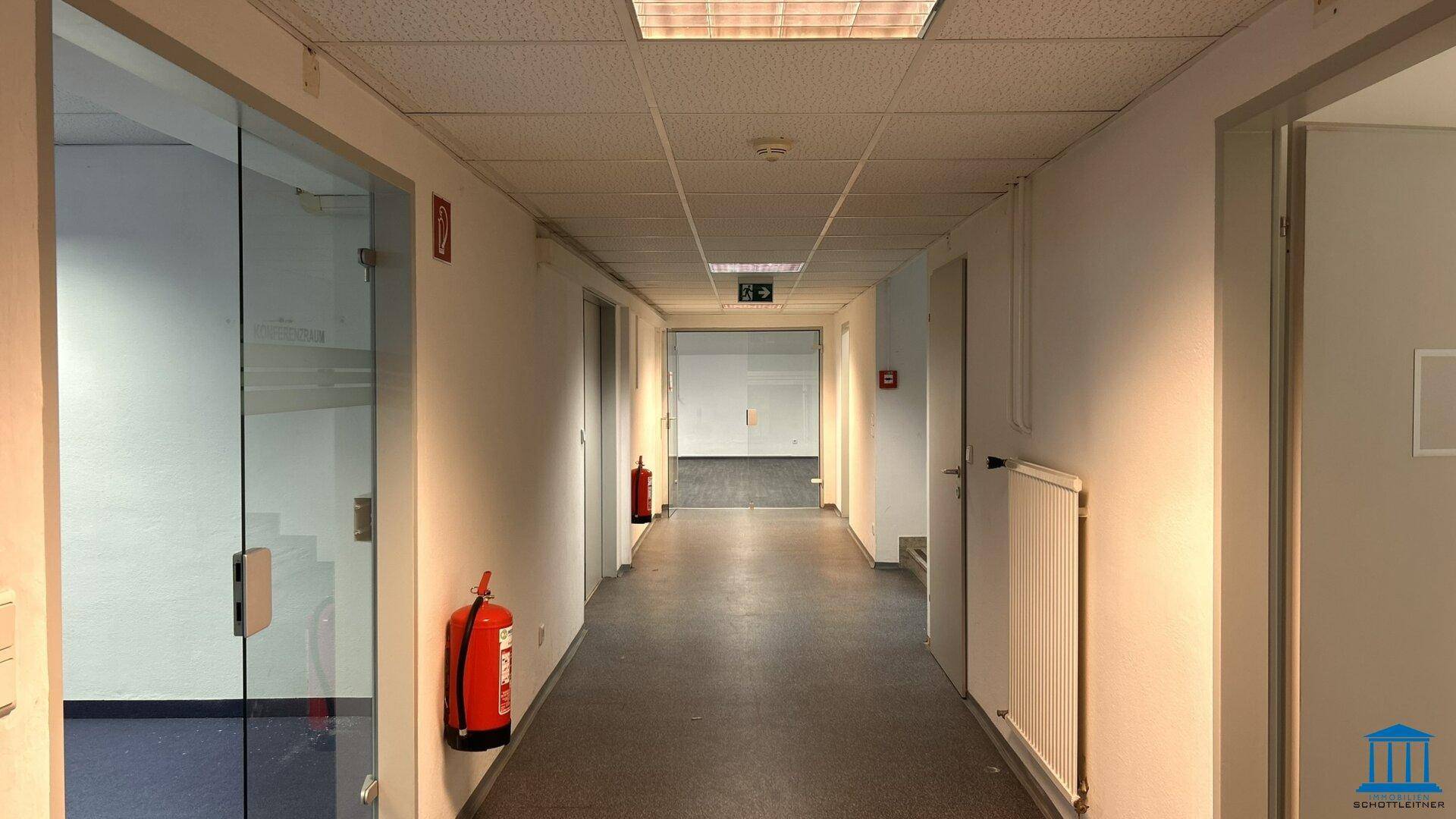 Langer Flur mit Teppichboden, Glastüren zu Büros und Brandschutzvorrichtungen.
