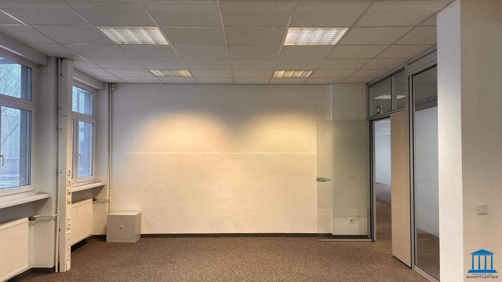 Heller Büroraum mit Teppichboden, mehreren Fenstern und einer Glastür.