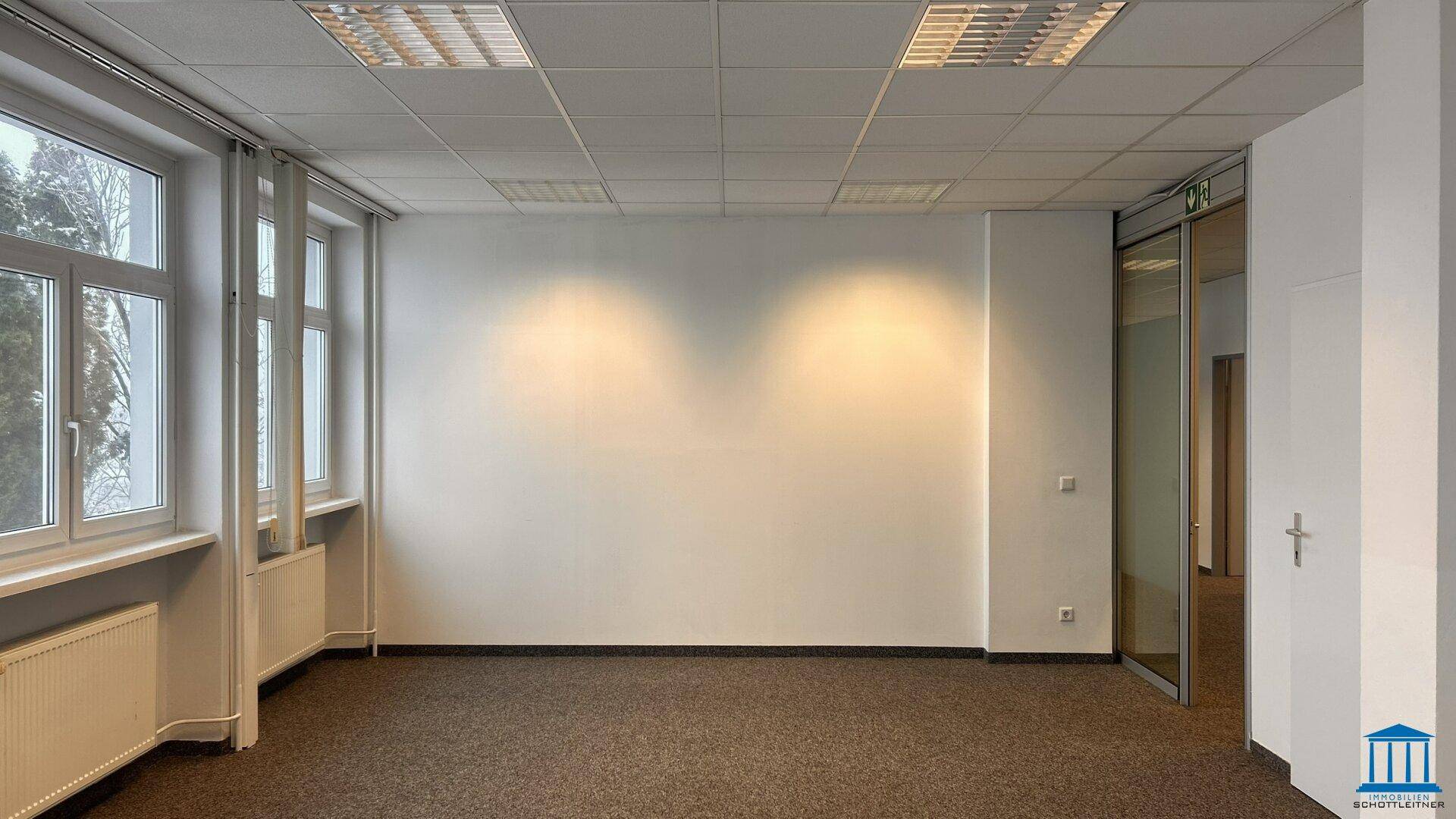 Helles, leeres Büro mit Teppichboden, großen Fenstern und einer Glaszugangstür, bereit für neue Mieter.
