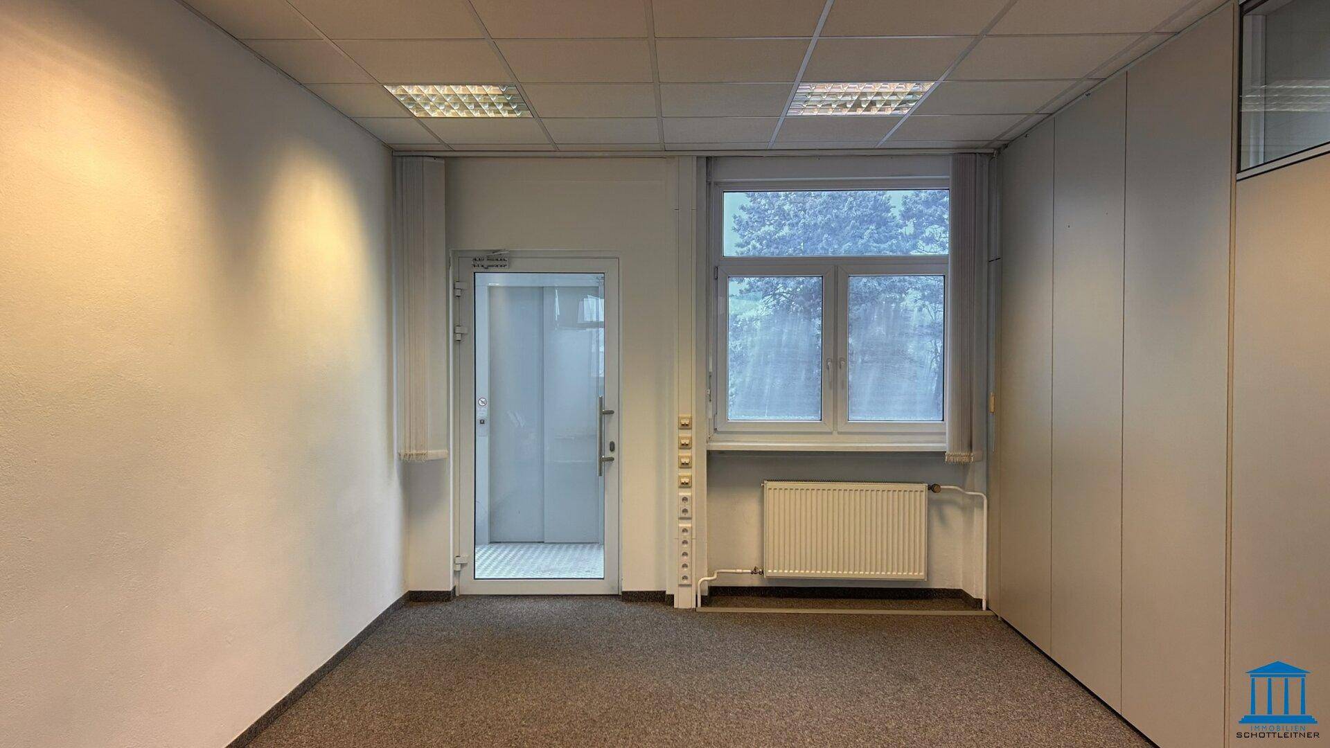 Büroraum mit Glasaufzug, Fenstern und integrierten Schränken, bietet eine praktische und helle Arbeitsumgebung.