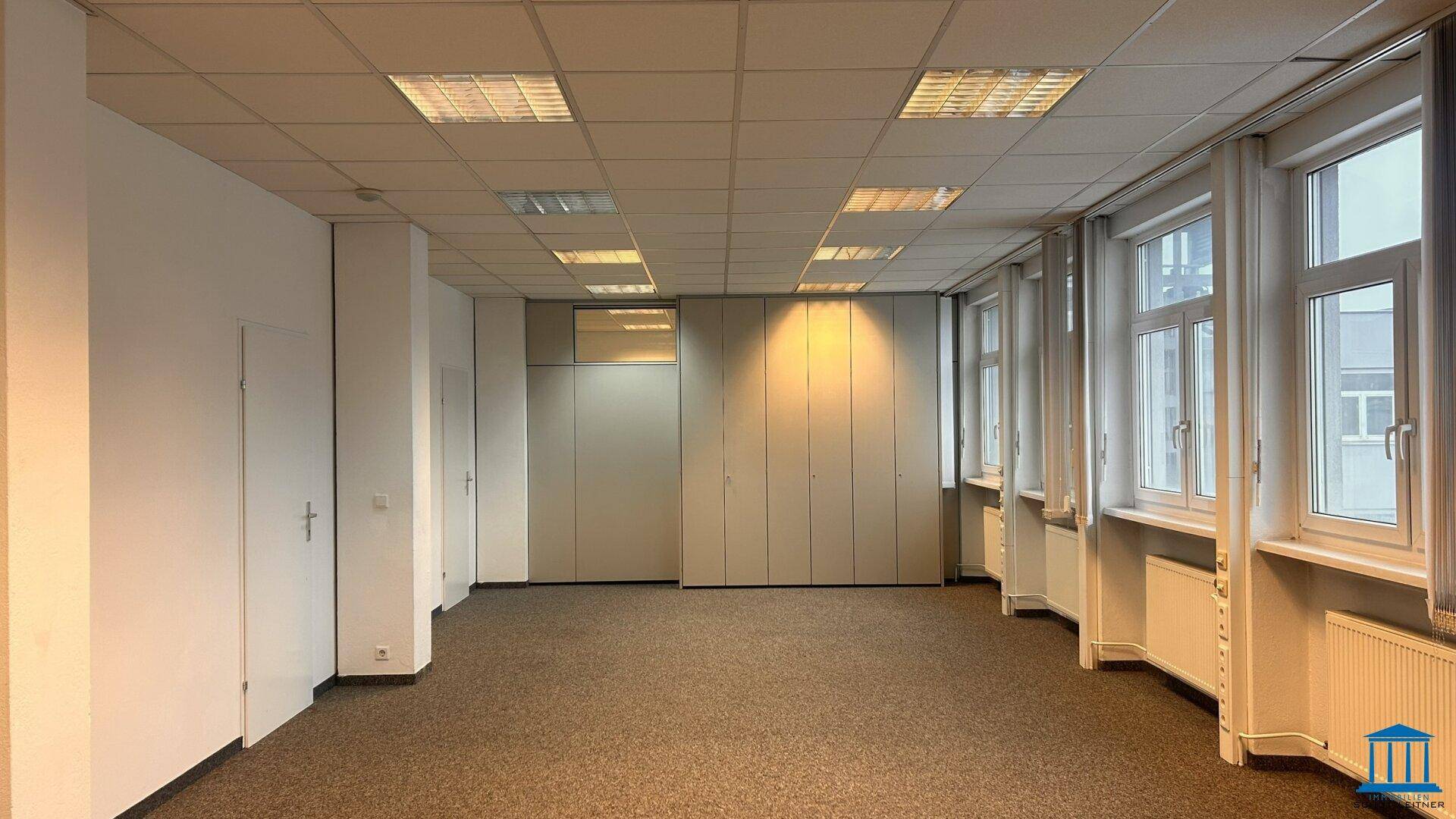 Geräumiges Büro mit Teppichboden, Fenstern und integrierten Schränken, bietet viel Stauraum und eine klare Struktur.