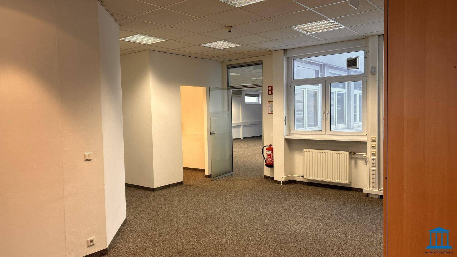 Helles Bürozimmer mit Teppichboden, großen Fenstern und einem Feuerlöscher, bereit für neue Mieter.
