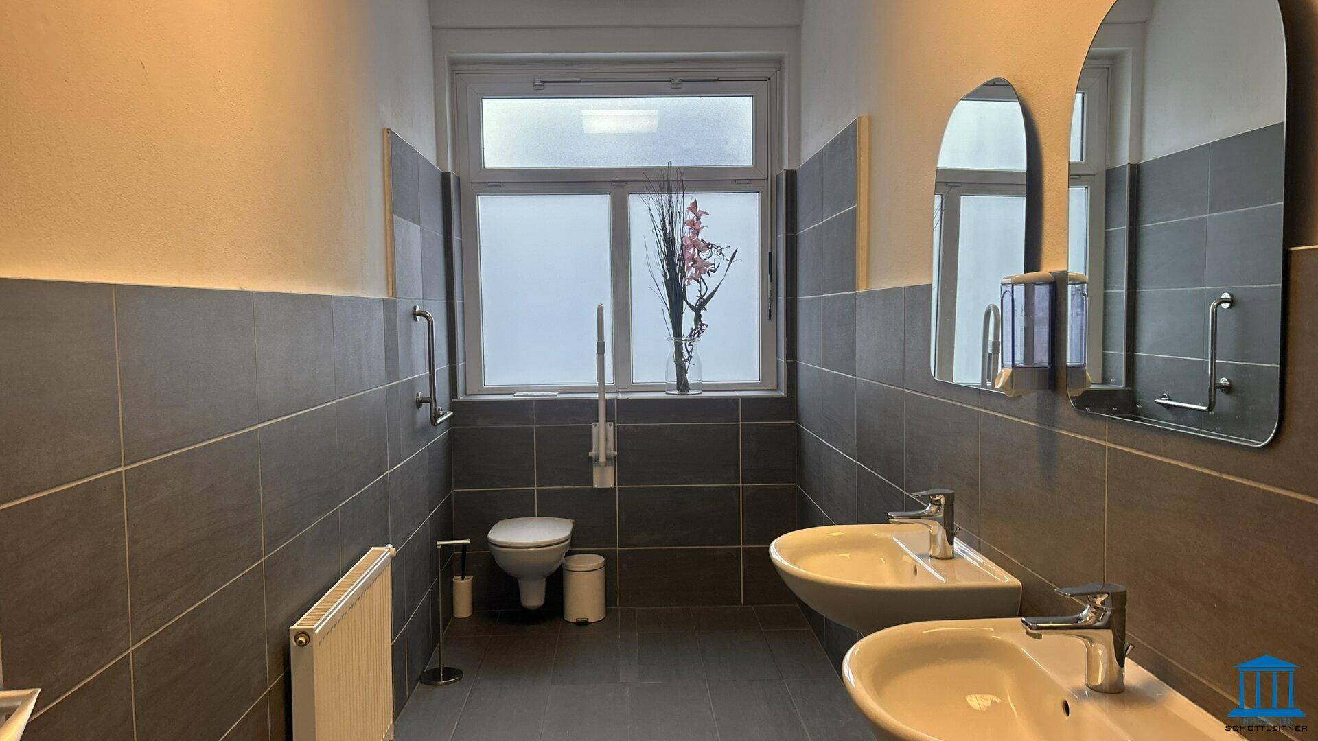 Geräumiges Badezimmer mit zwei Waschbecken, Toilette, Haltegriffen und Fenster, modern gefliest.