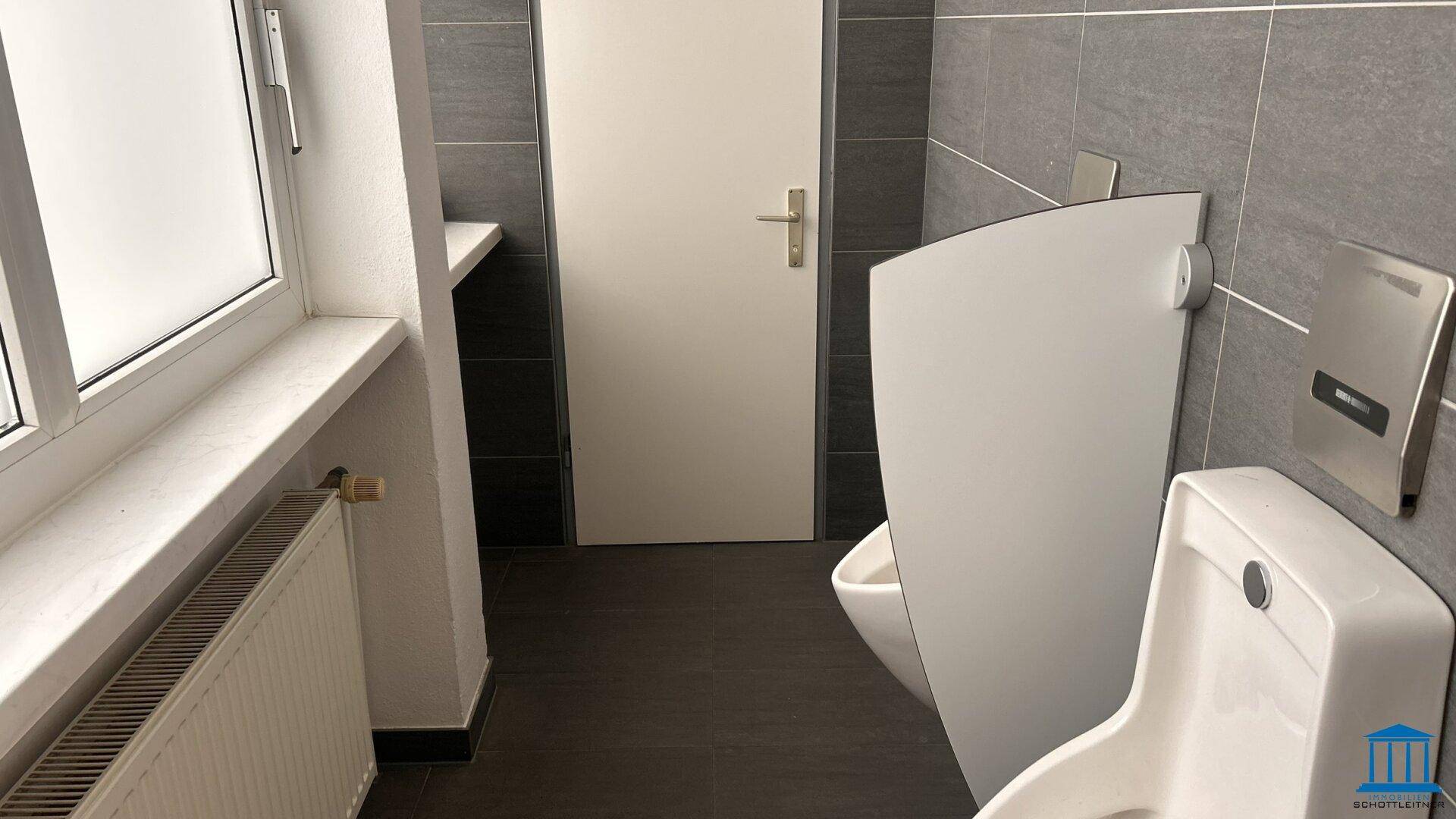 Sanitärbereich mit Urinal, Toilette und Fenster, ausgestattet mit dunklen Fliesen und Heizkörper.