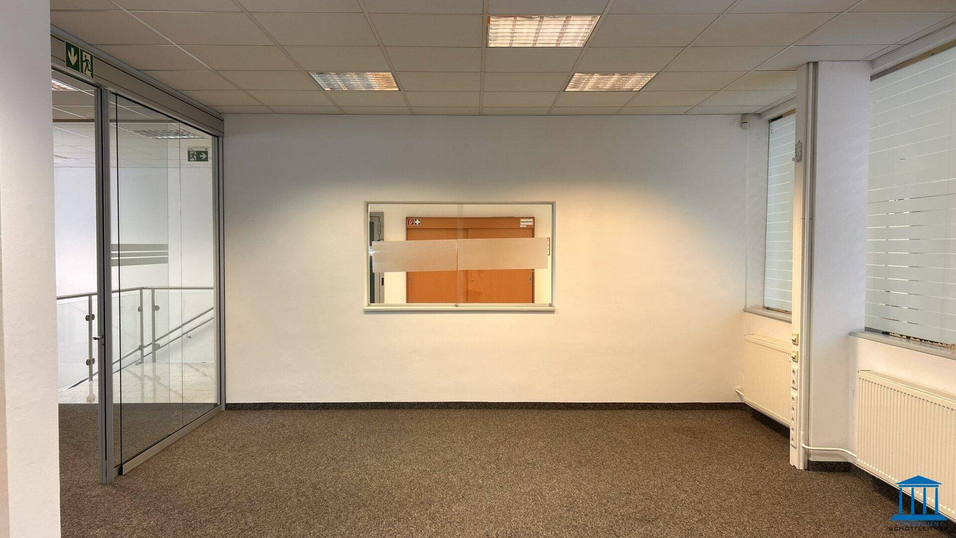 Leeres Büro mit Teppichboden, einem Fenster und einer Glaswand, die den Raum abgrenzt.