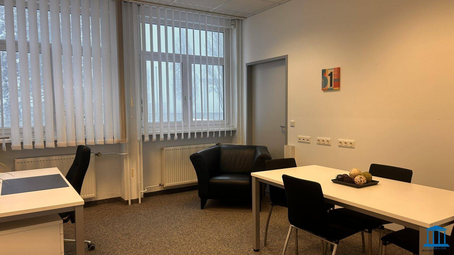 Möbliertes Büro mit Teppichboden, Schreibtisch, Sessel und einem Tisch mit Stühlen.