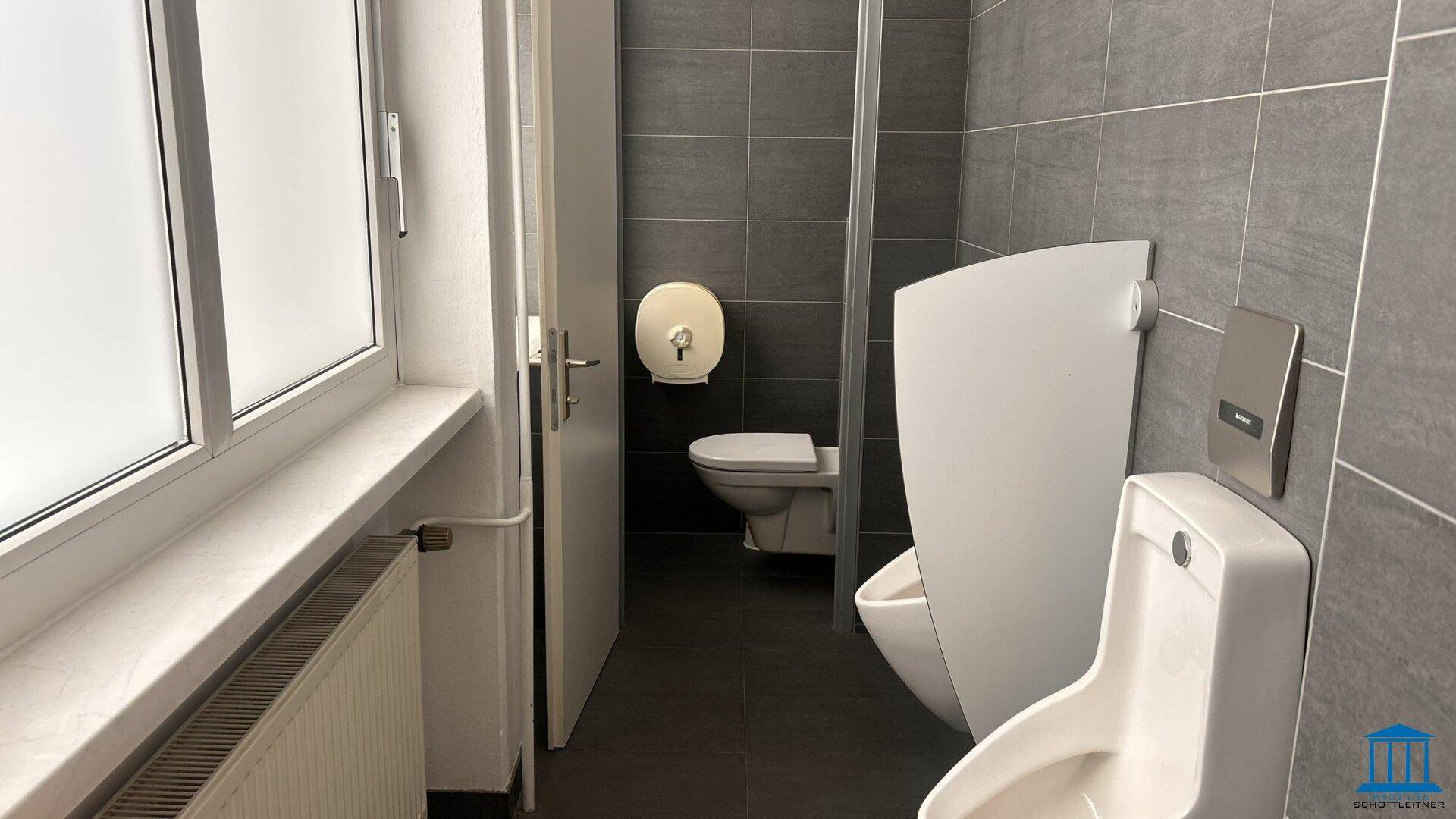 Modernes Badezimmer mit grauen Fliesen, Urinal und Toilette, gut beleuchtet.