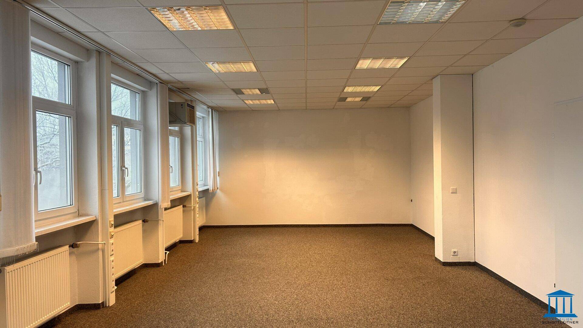 Großes, leeres Büro mit vielen Fenstern, Teppichboden und Rasterdeckenbeleuchtung.