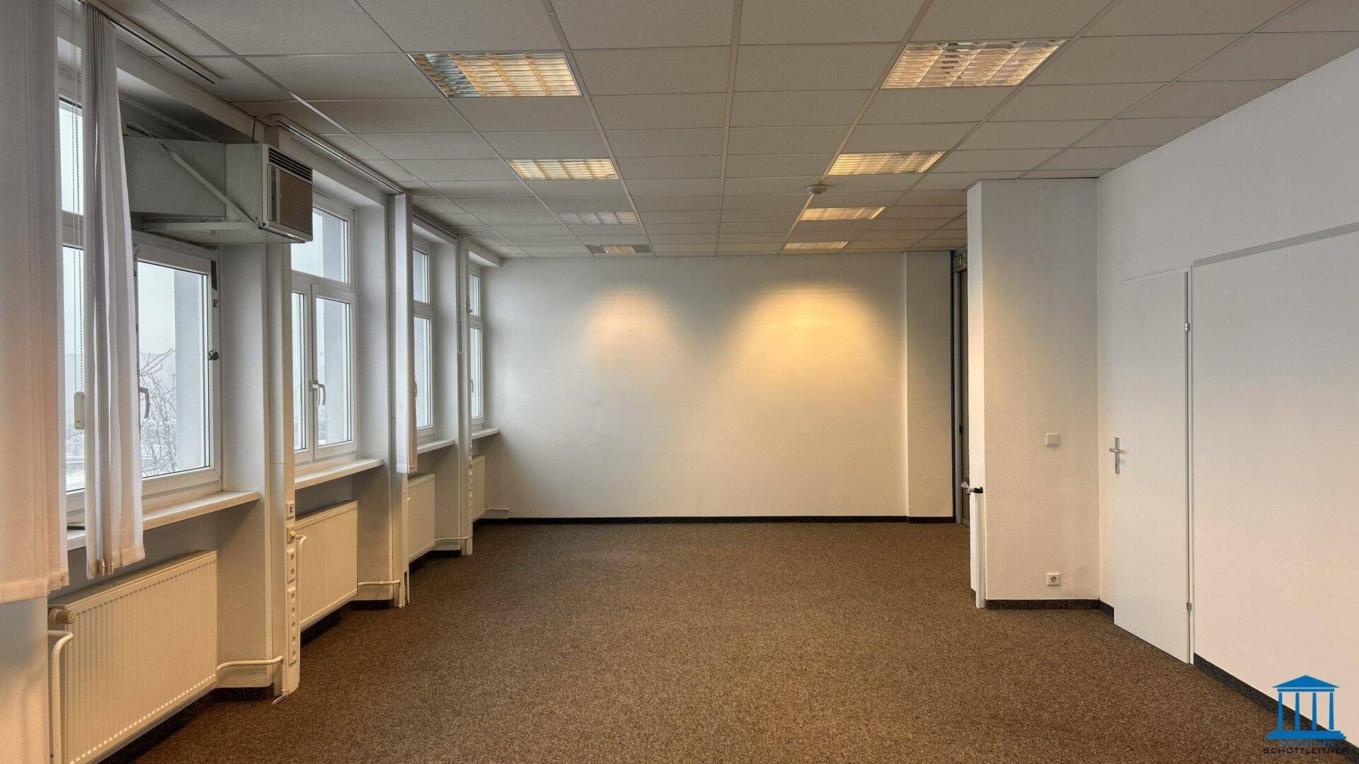 Helles Büro mit Teppichboden, weißen Wänden und mehreren Fenstern, die viel Tageslicht hereinlassen.
