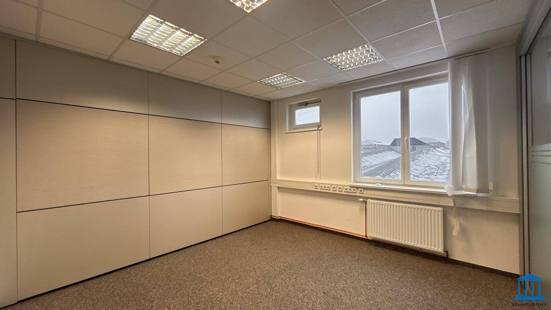 Büro mit Teppichboden, Trennwänden und einem Fenster mit Blick nach draußen.