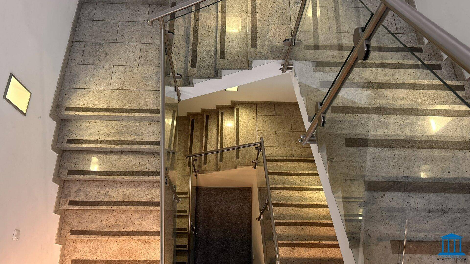 Modernes Treppenhaus mit Steinfliesen, Glasgeländer und Edelstahlhandläufen, gut beleuchtet durch indirektes Licht.