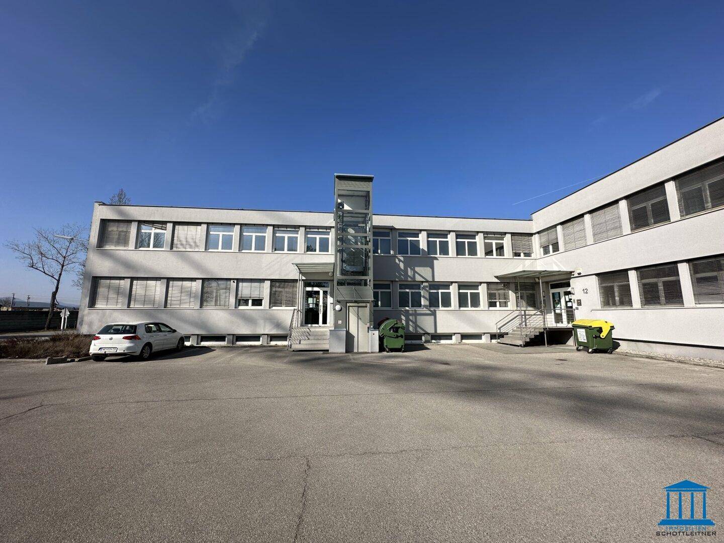 Vorderansicht eines Bürogebäudes mit Eingangsbereich, Parkplätzen und einem externen Glasaufzug.