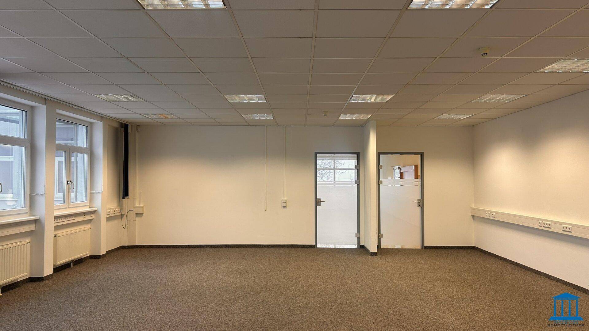 Helles, leeres Büro mit Teppichboden und zwei Türen, bereit zur individuellen Gestaltung.