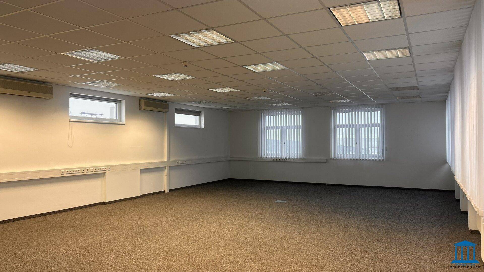 Großes, leeres Büro mit Teppichboden, Fenstern und Klimaanlage.