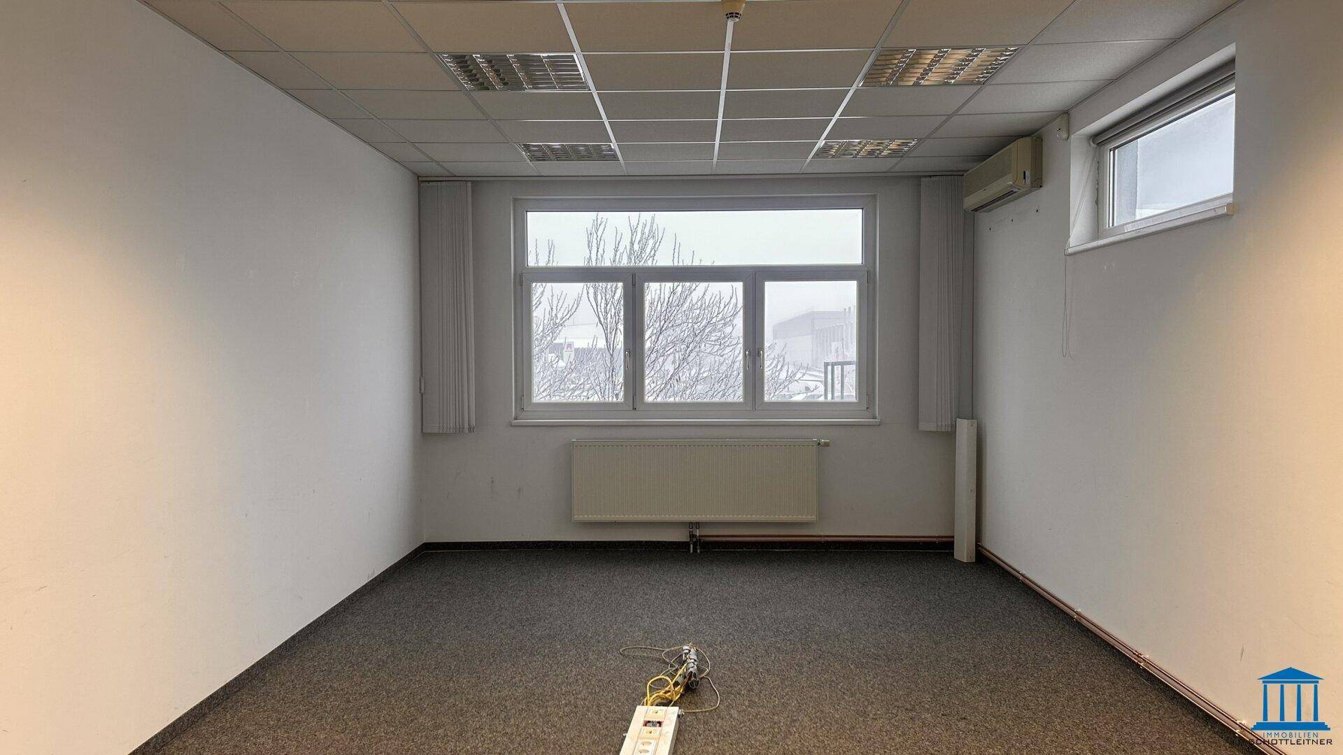 Helles Büro mit Teppichboden, großem Fenster und Heizkörper, bietet eine angenehme Arbeitsumgebung.