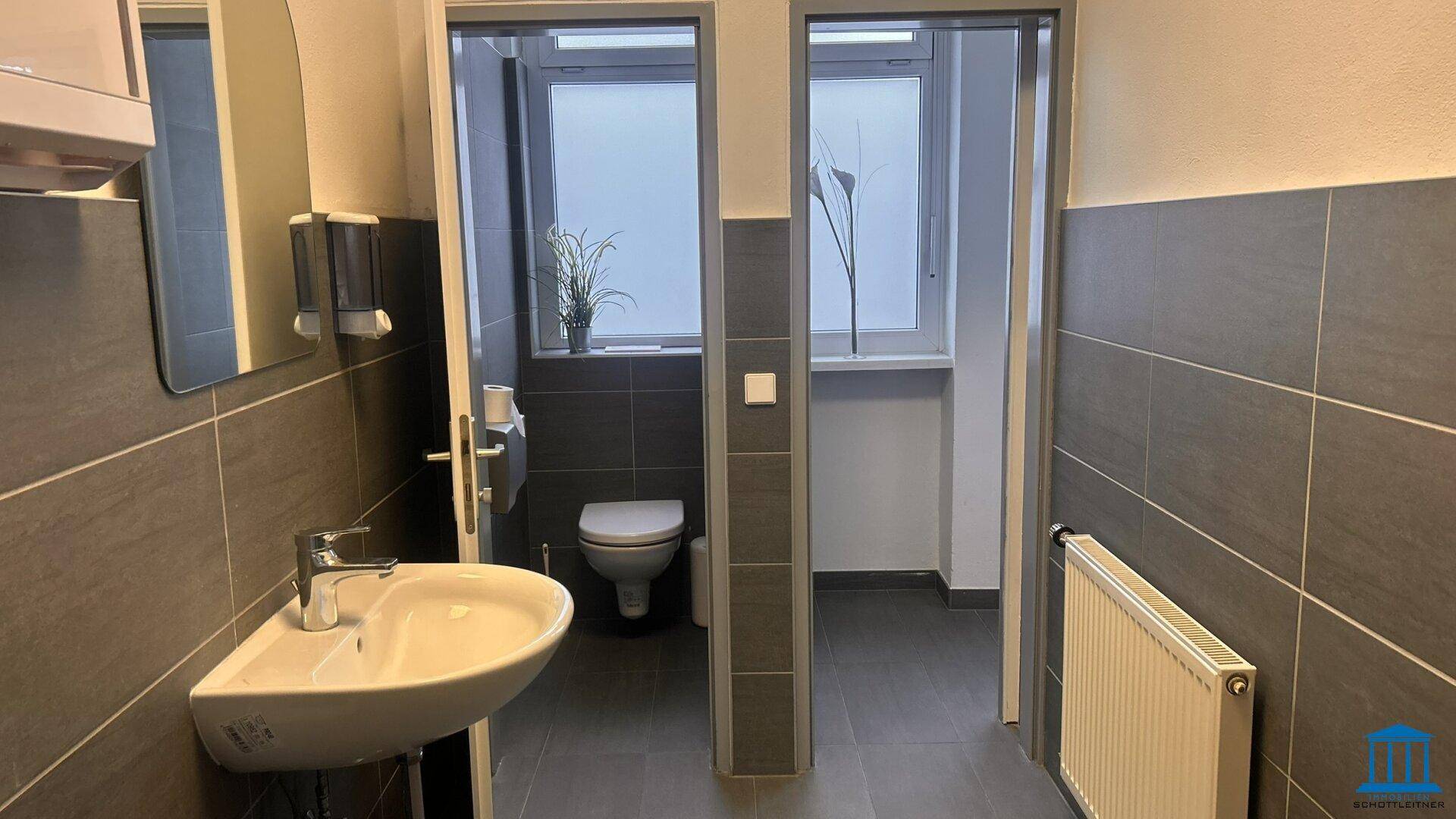 Badezimmer mit Waschbecken, Wand-WC und Fenster, getrennt durch eine Tür zu einem weiteren WC-Bereich.