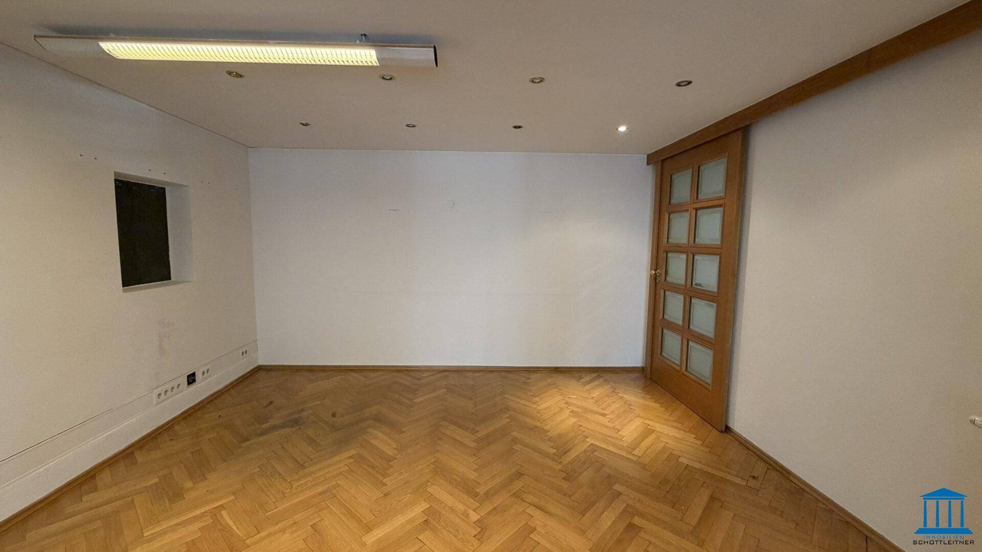 Leerer Büroraum mit klassischem Fischgrätparkett, weißen Wänden und einer Holztür mit Glaseinsätzen.