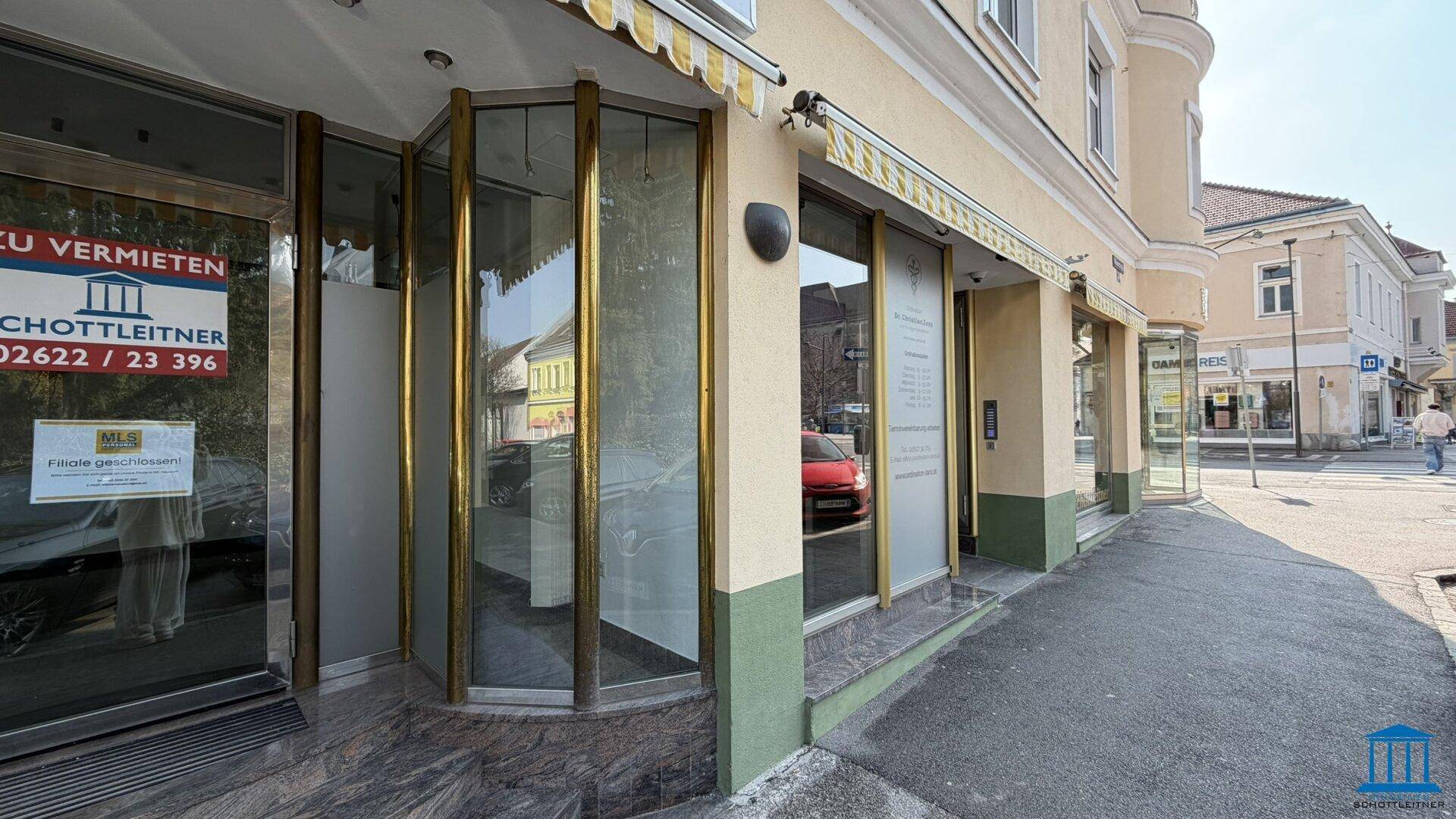 Eckgebäude mit großen Schaufenstern, Markisen und einer breiten Gehwegfläche vor dem Eingang.