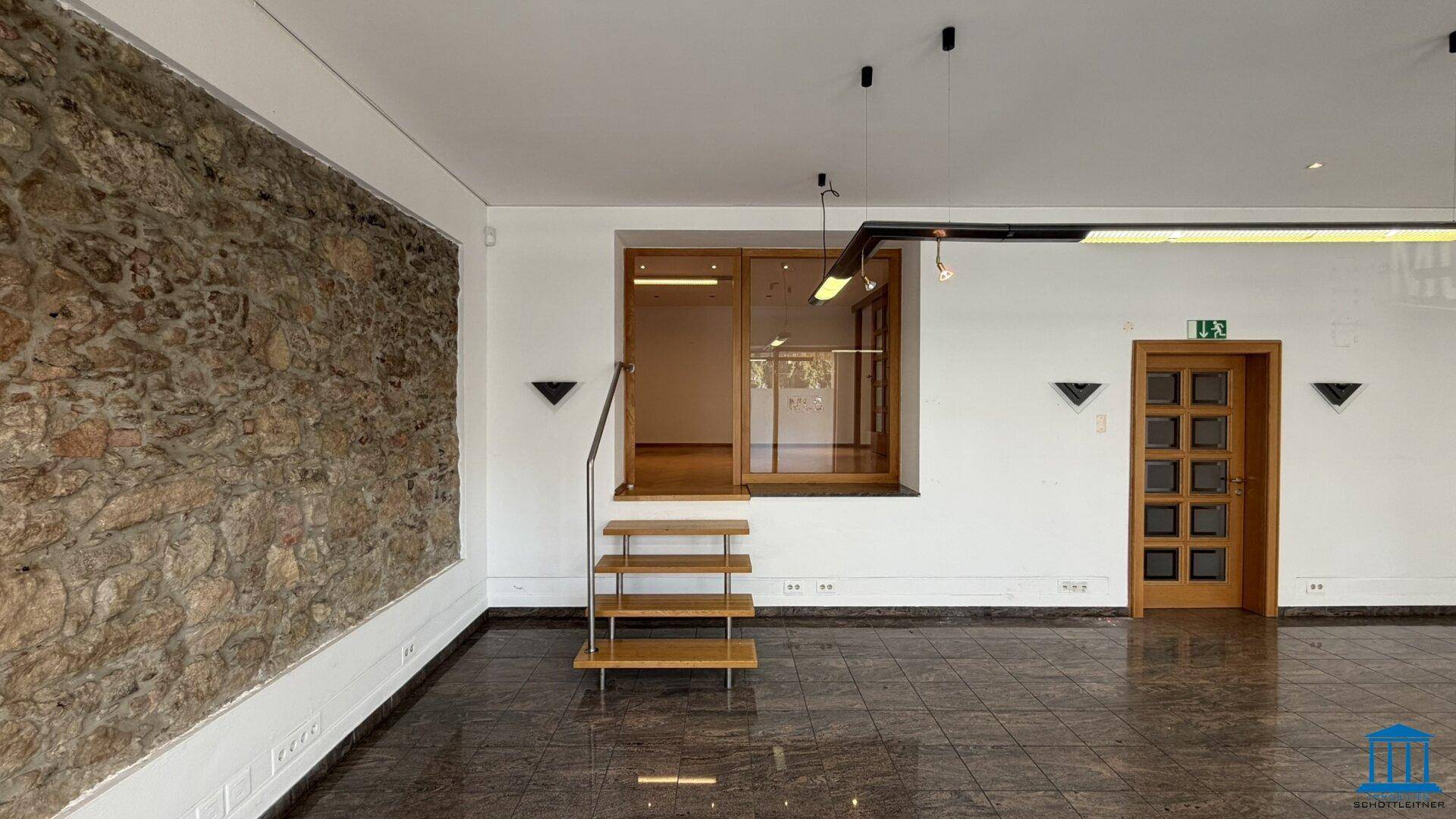 Gewerberaum mit markanter Steinwand, glänzendem Fliesenboden und einer kleinen Holztreppe zu einem erhöhten Bereich.