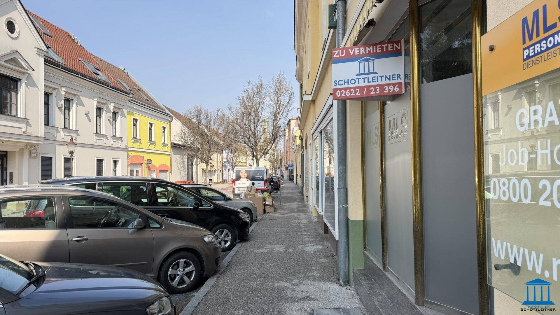 Straßenansicht mit parkenden Autos und der Geschäftsfassade mit "Zu vermieten"-Schildern unter blauem Himmel.