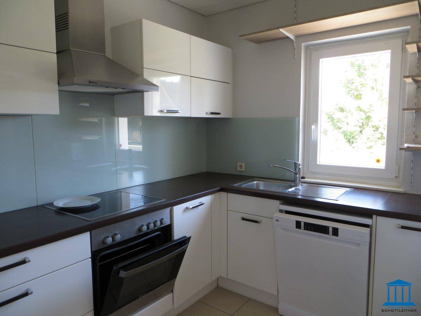 Helle moderne Küche mit weißen Hochglanzfronten, dunkler Arbeitsplatte und Fenster für Tageslicht.
