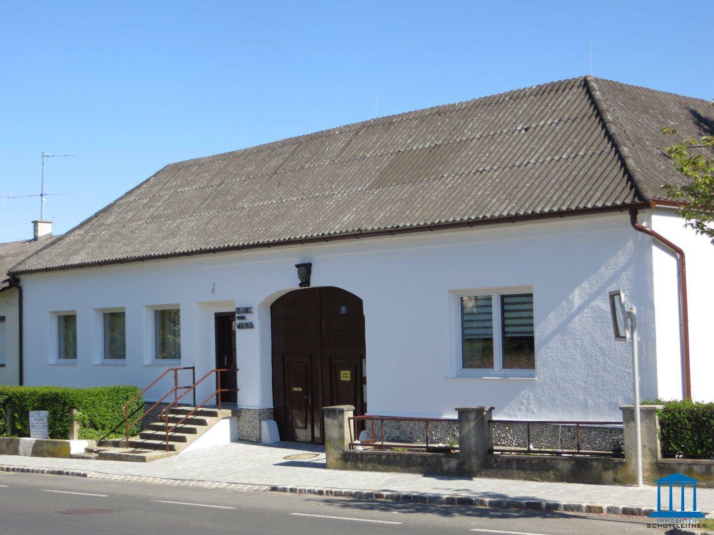 Ansicht des Nebengebäudes mit weißer Fassade, dunklem Satteldach und gepflegter Hecke.