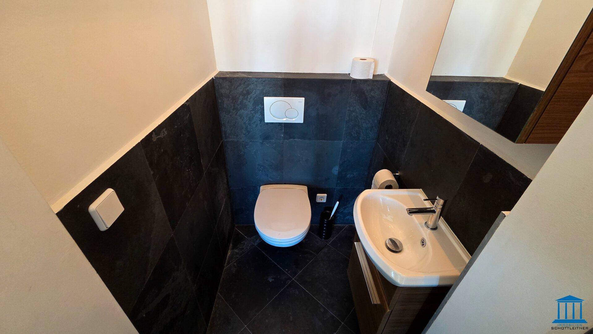 Kleines Gäste-WC mit Wand-WC, Waschbecken und dunklen Fliesen.