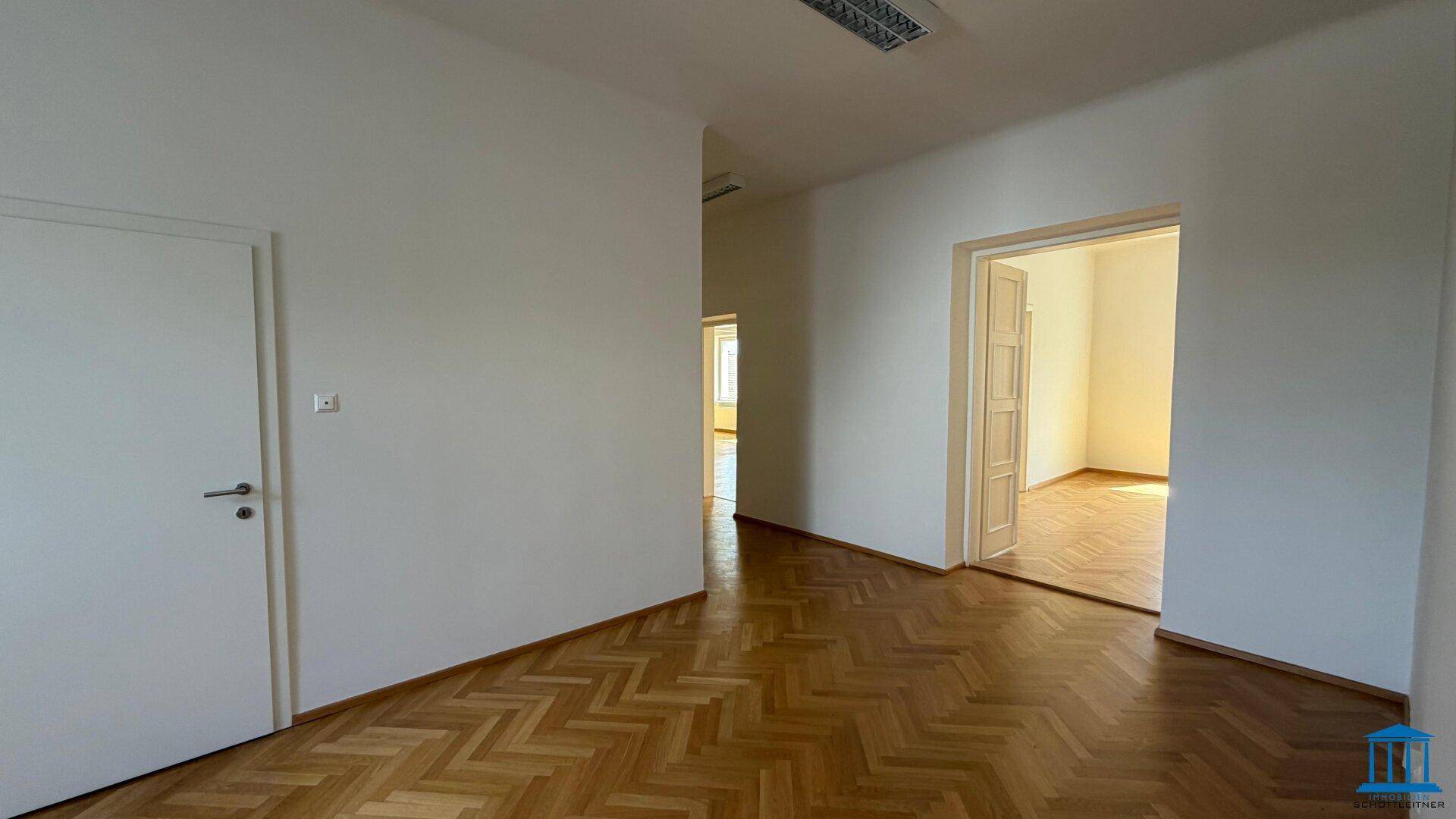 Leeres Zimmer mit Fischgrätparkett, einer geschlossenen Tür und einem offenen Durchgang zu einem anderen Bereich.