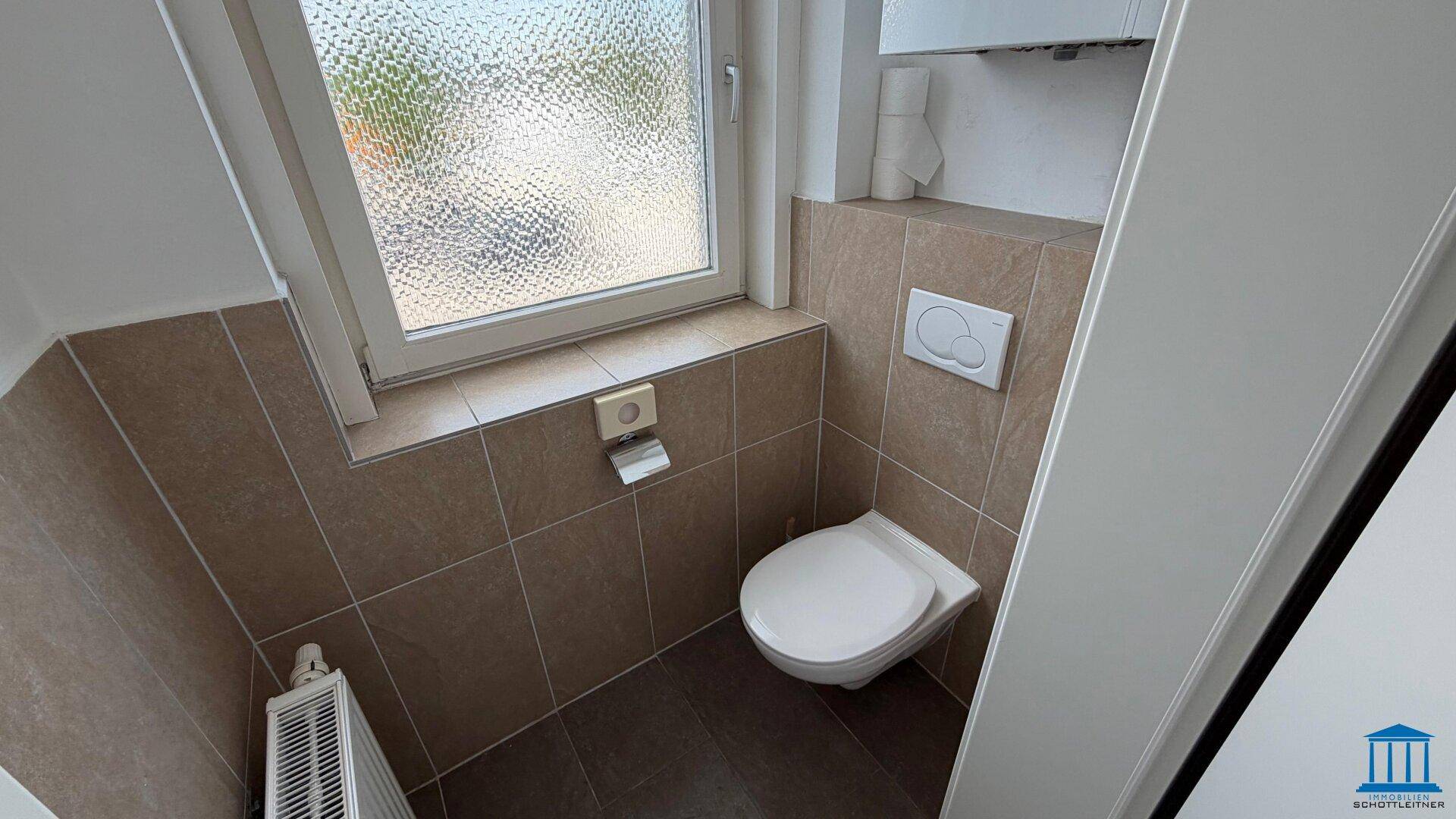 WC-Raum mit wandhängendem WC, einem Fenster und braunen Fliesen an den Wänden.