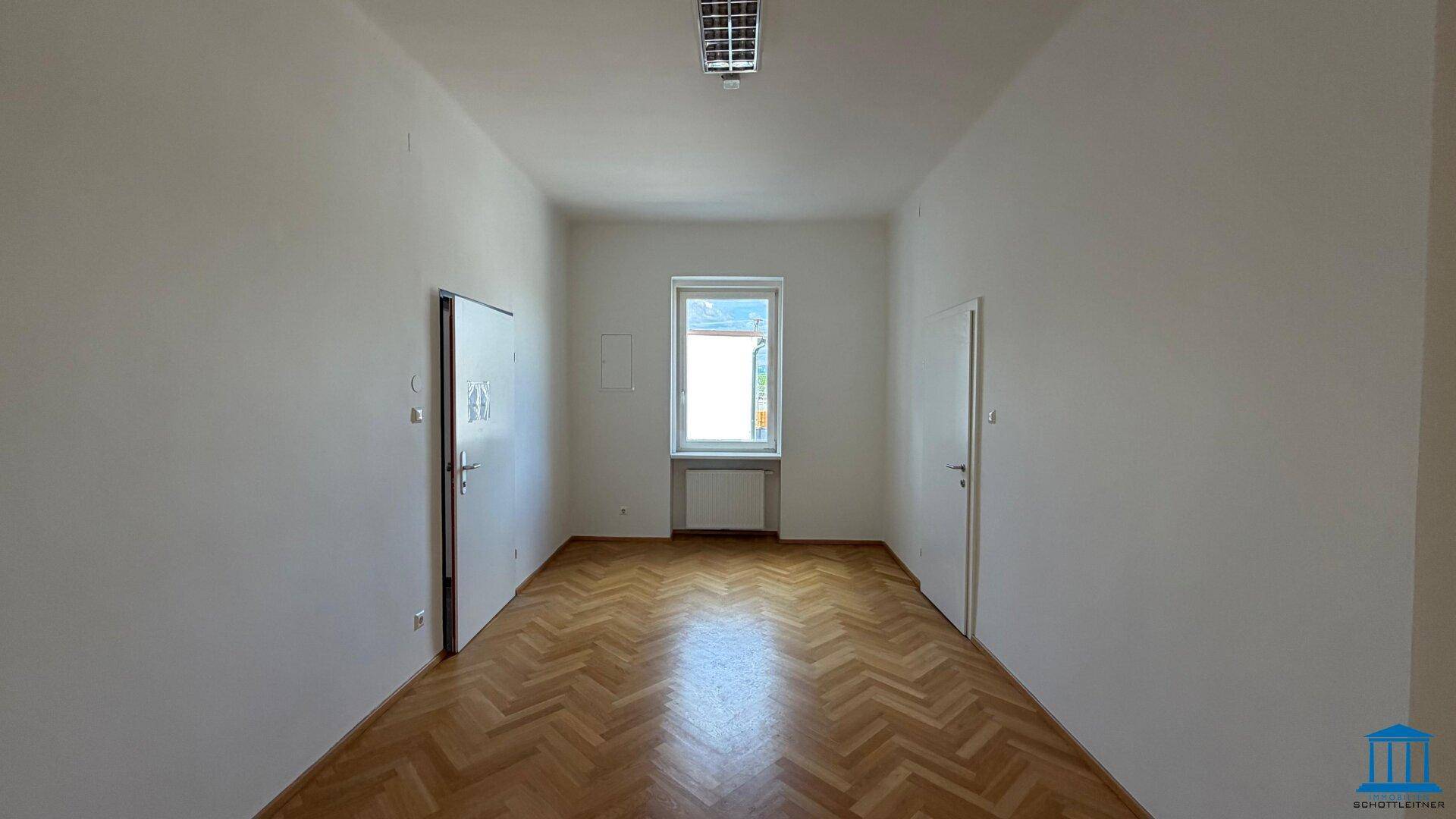 Helles, leeres Zimmer mit Fischgrätparkett, einem Fenster und Türen auf beiden Seiten.