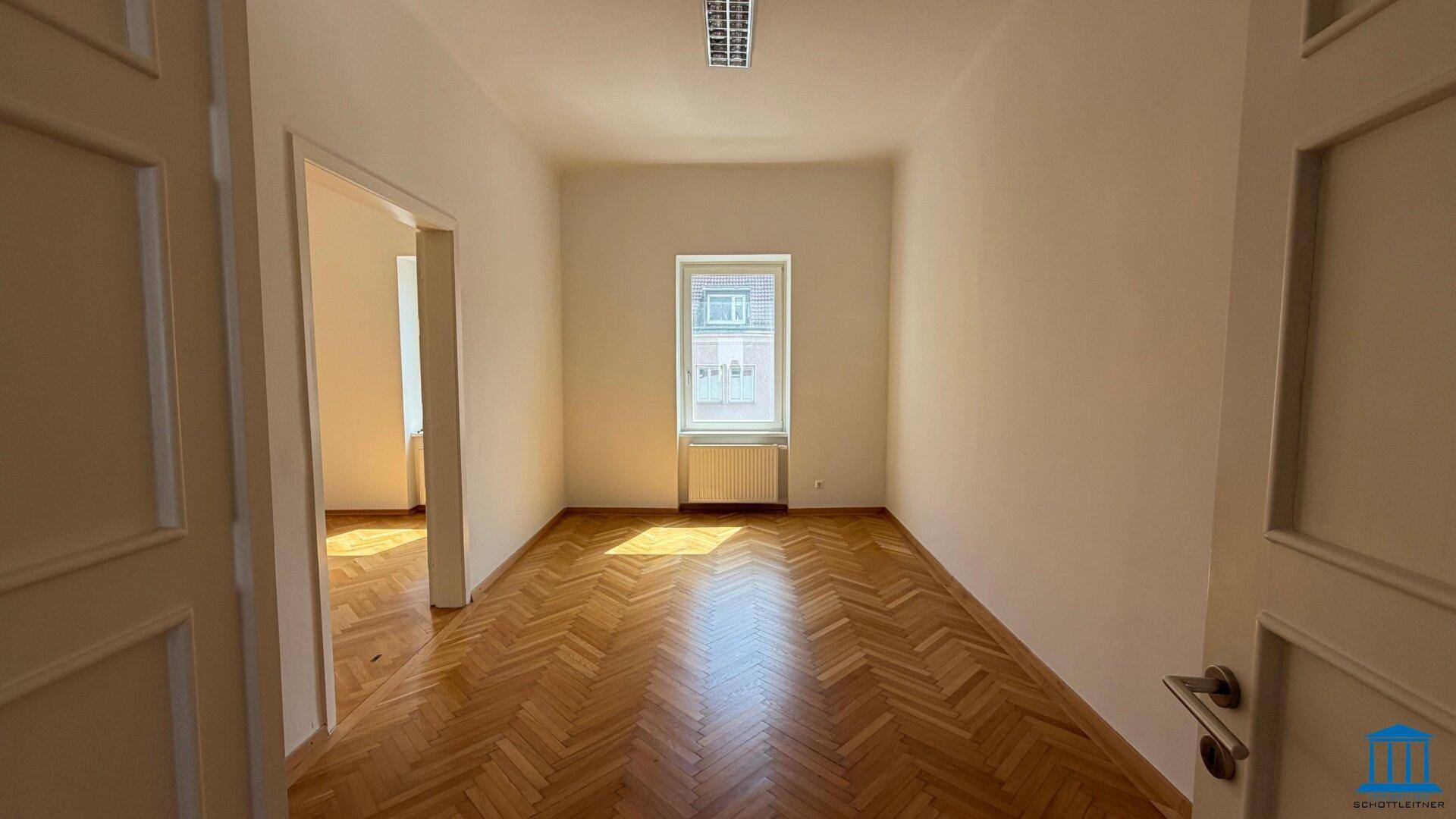 Helles, leeres Zimmer mit Fischgrätparkett, einem Fenster und einem Heizkörper darunter.